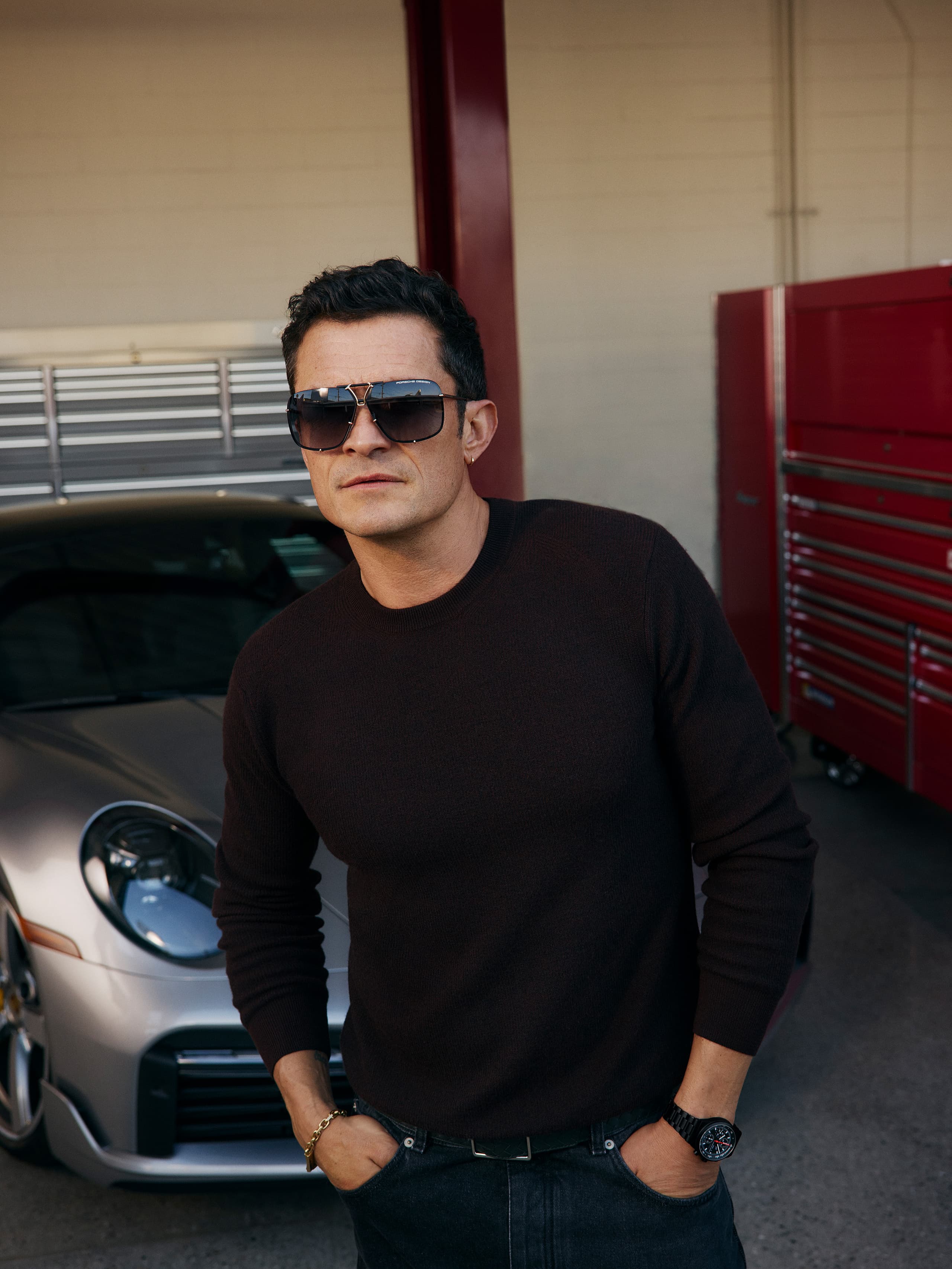 Man in sunglasses and dark sweater stands by a silver Porsche in a garage with red tool cabinets.