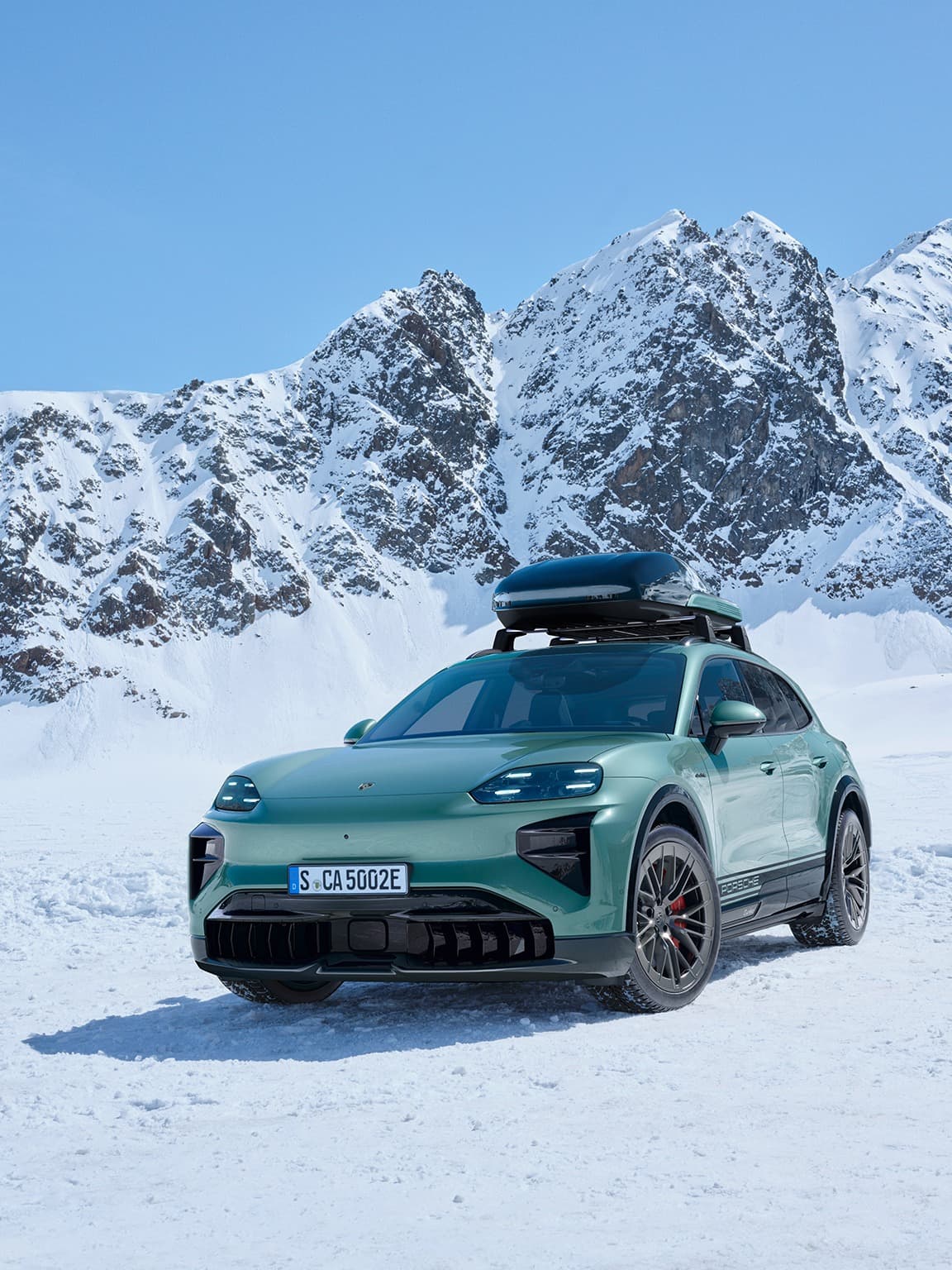 Green Porsche SUV with roof box on snowy terrain, set against a backdrop of snow-covered mountains under a clear blue sky.