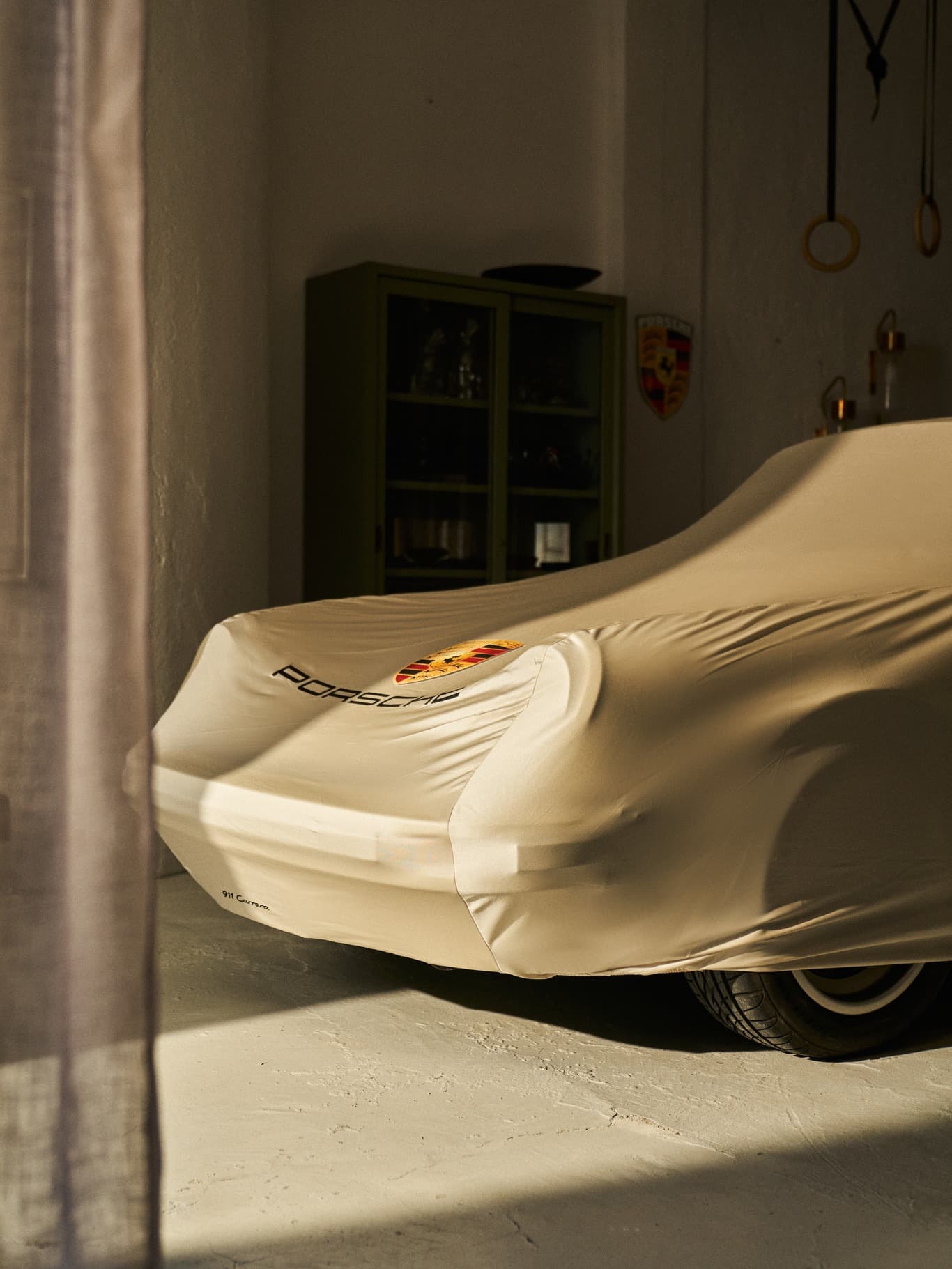 Porsche 911 Carrera under beige cover in a dimly lit garage, with a cabinet and Porsche emblem in the background.