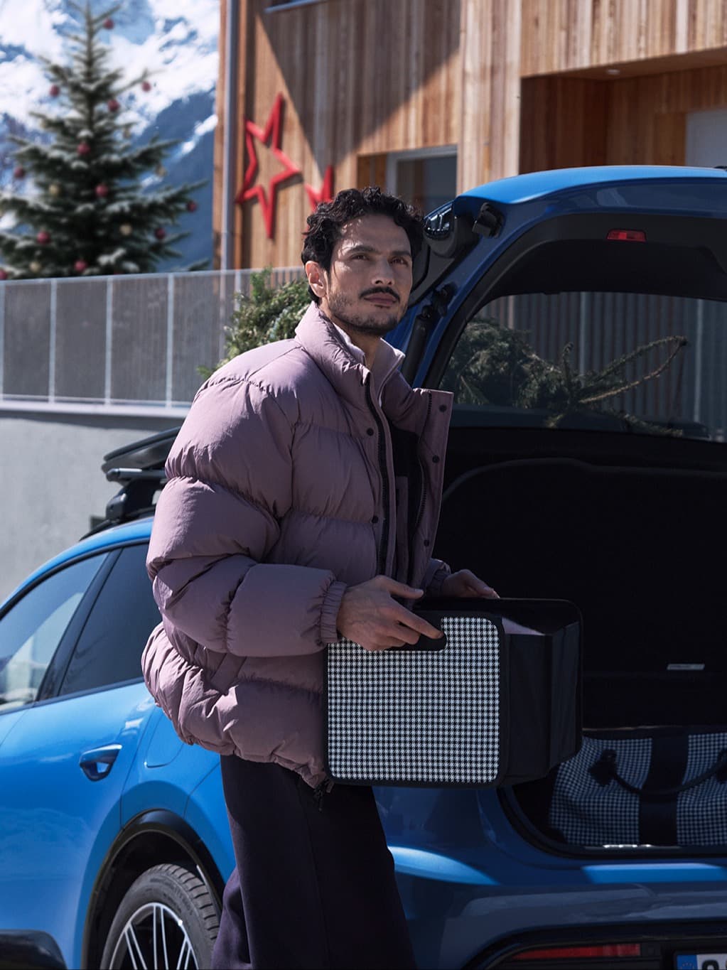 Man in a purple jacket holding a box by a blue car, with a snowy mountain and decorated tree in the background.