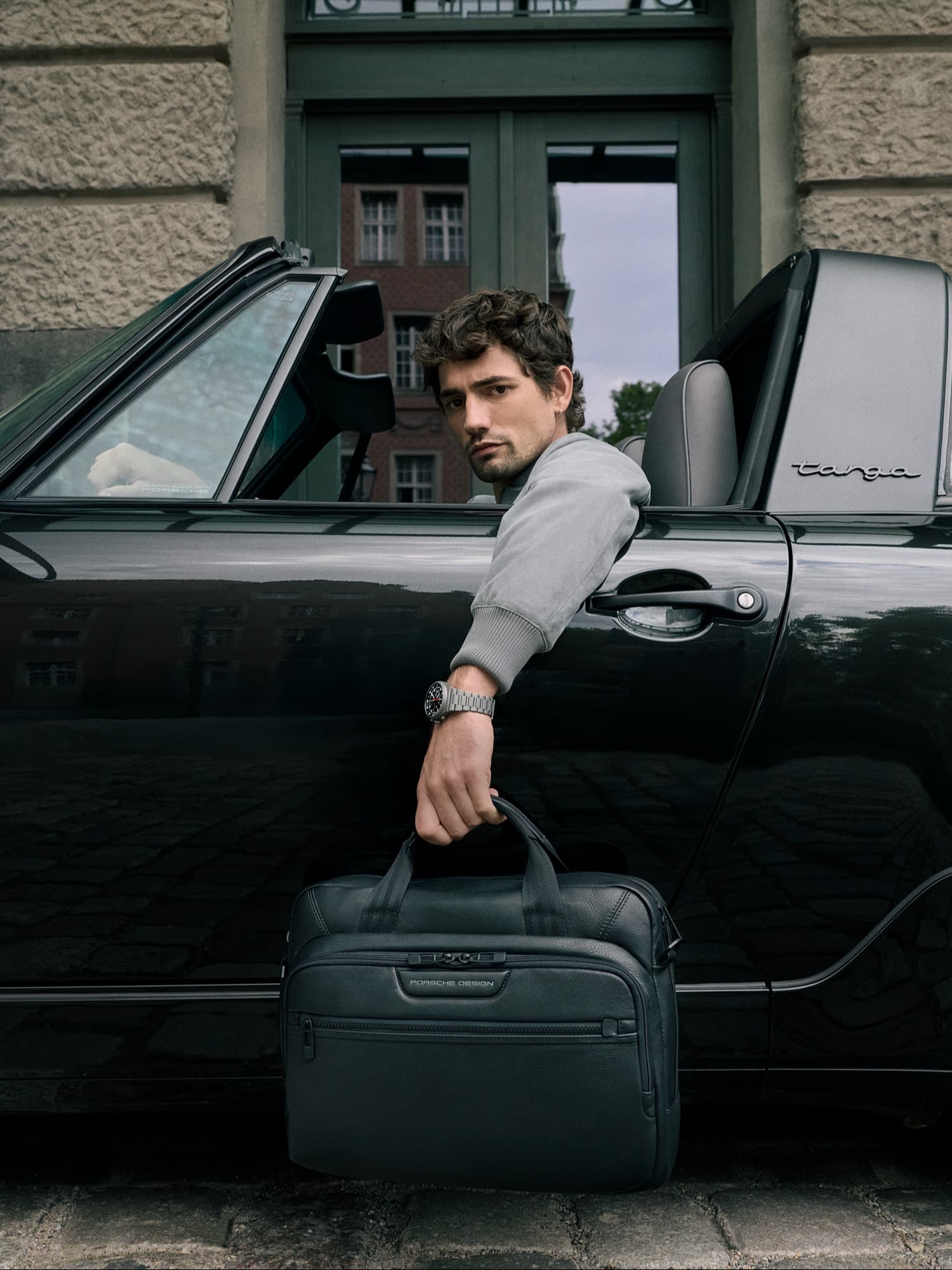 Mann im Auto hält eine schwarze Porsche Design Tasche, trägt eine Uhr, mit einem Stadtgebäude im Hintergrund.