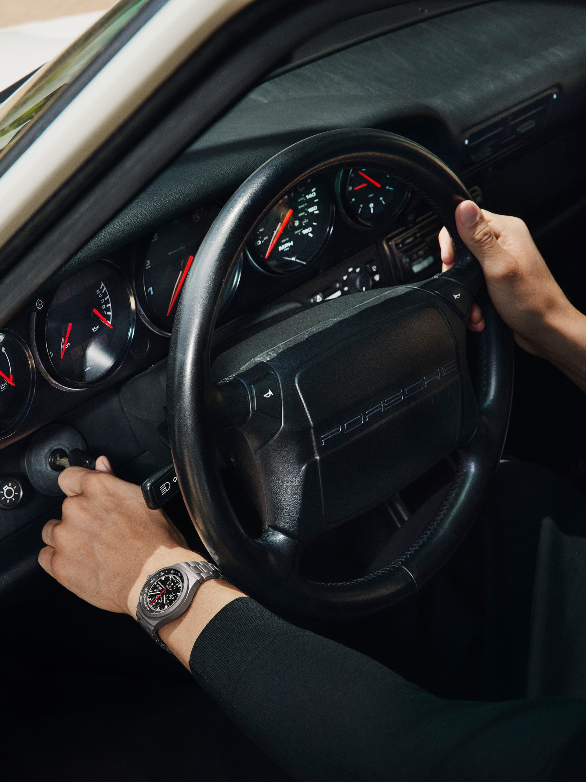 Hände am Porsche-Lenkrad, Armaturenbrett-Anzeigen sichtbar, Person trägt silberne Uhr, Autoinnenraum in Schwarz