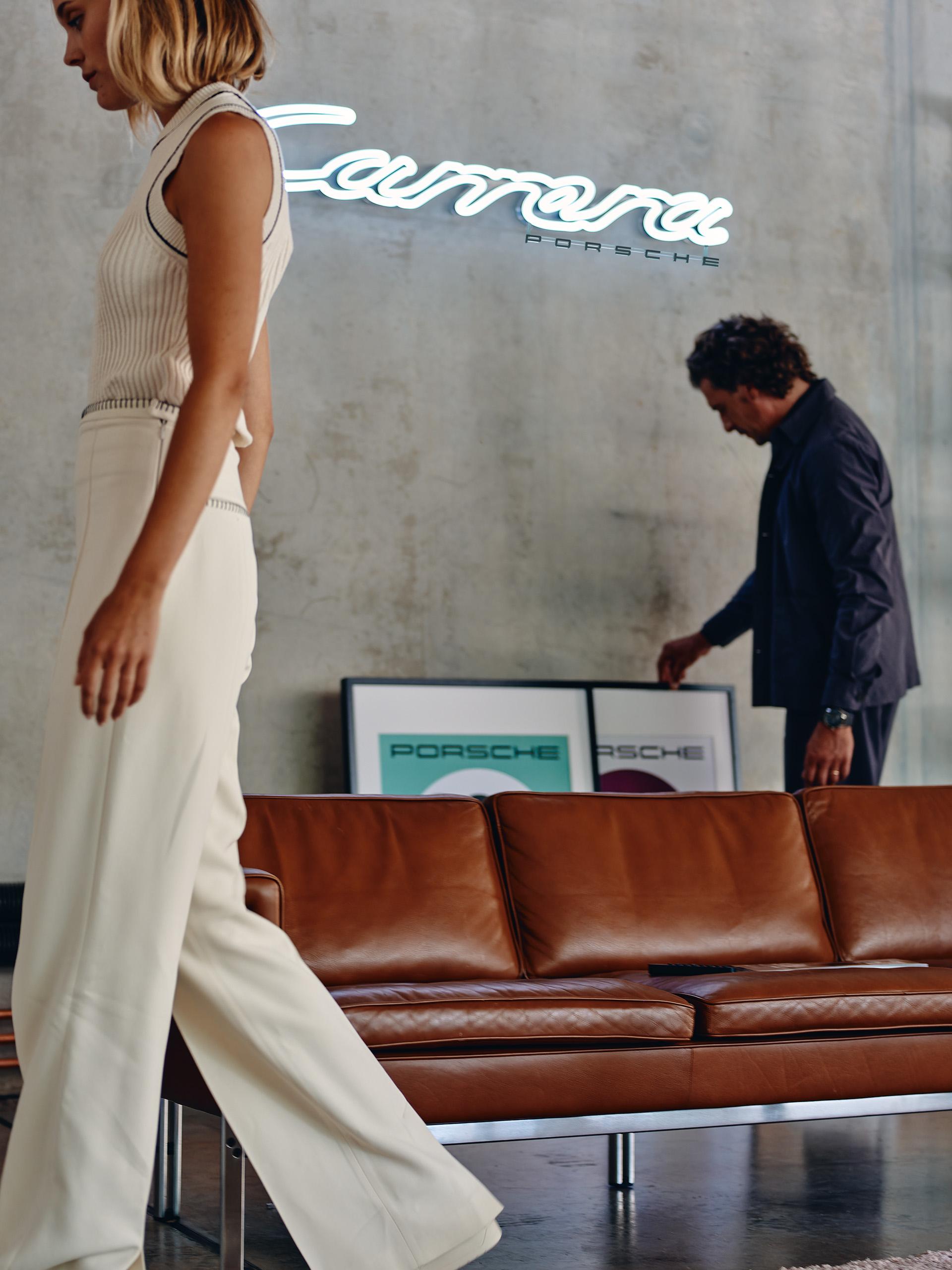 The picture shows a man and a woman in a room. The woman walks by and the man examines the pictures standing on the wall. A white Carrera typewriter is on the wall and a brown leather couch is in the foreground.