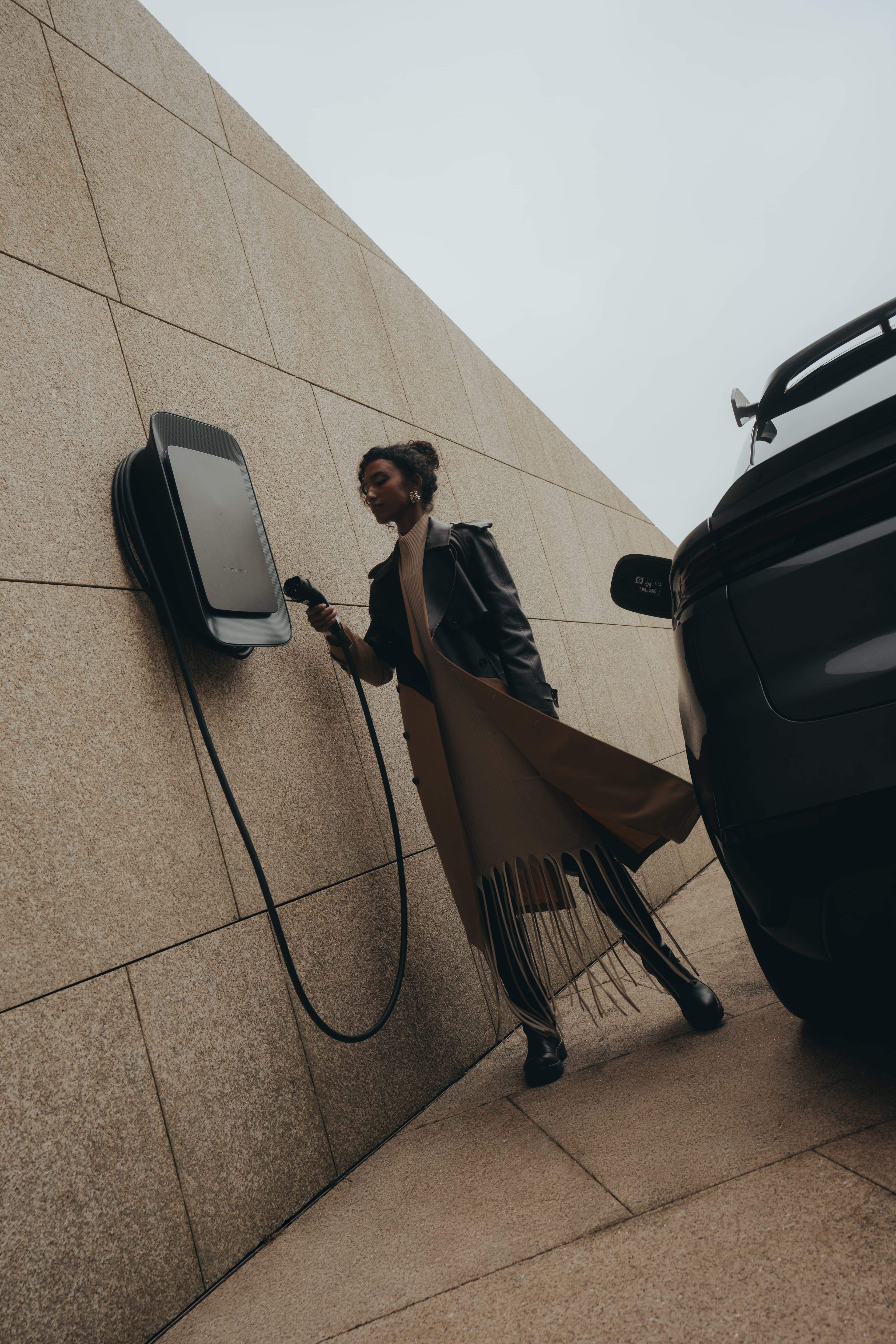The image shows a woman charging an electric car at a Porsche Wallbox mounted on a stone wall