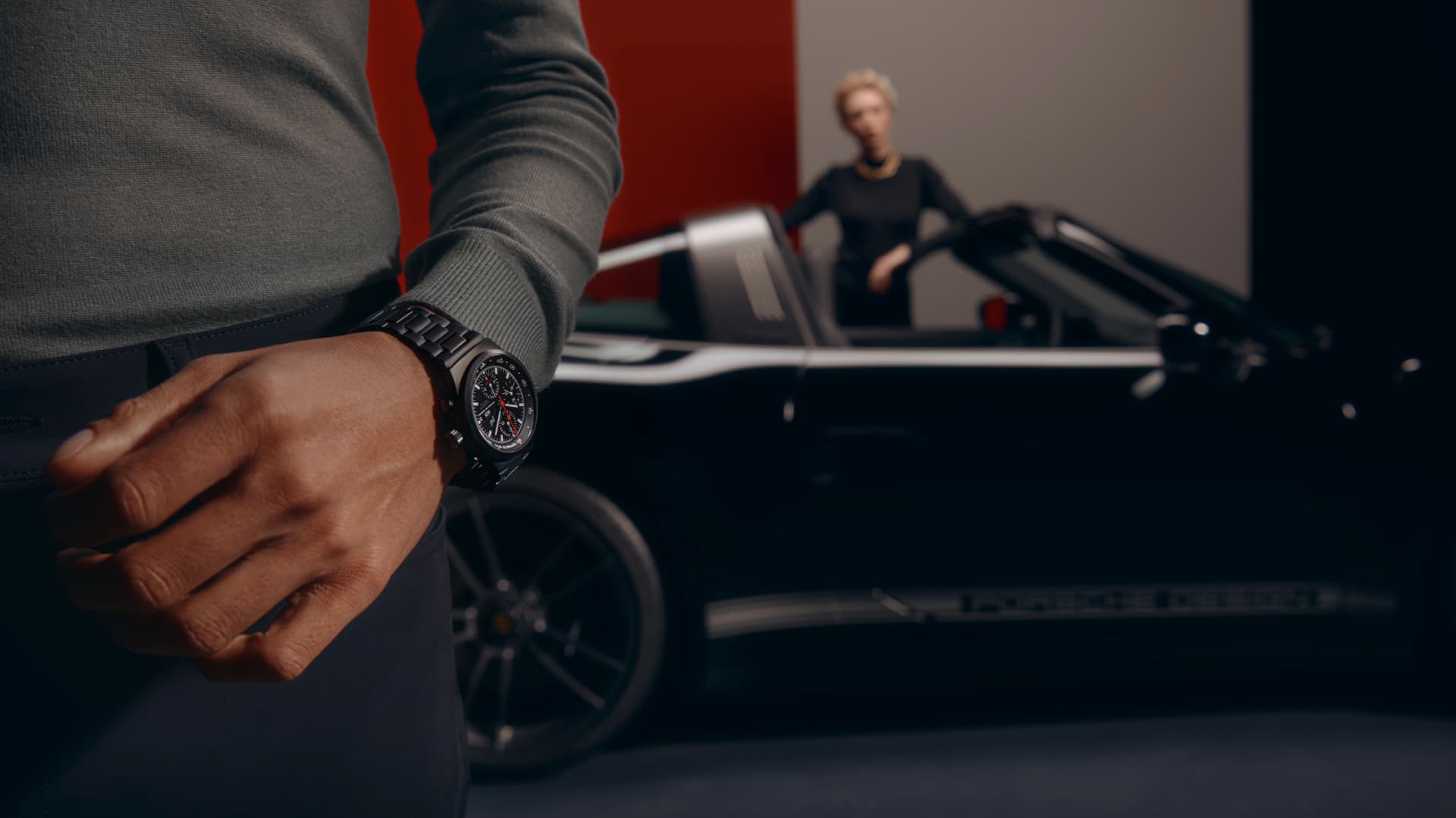 The picture shows a man wearing a black Porsche wristwatch and in the background is a black 911, also with a man in it