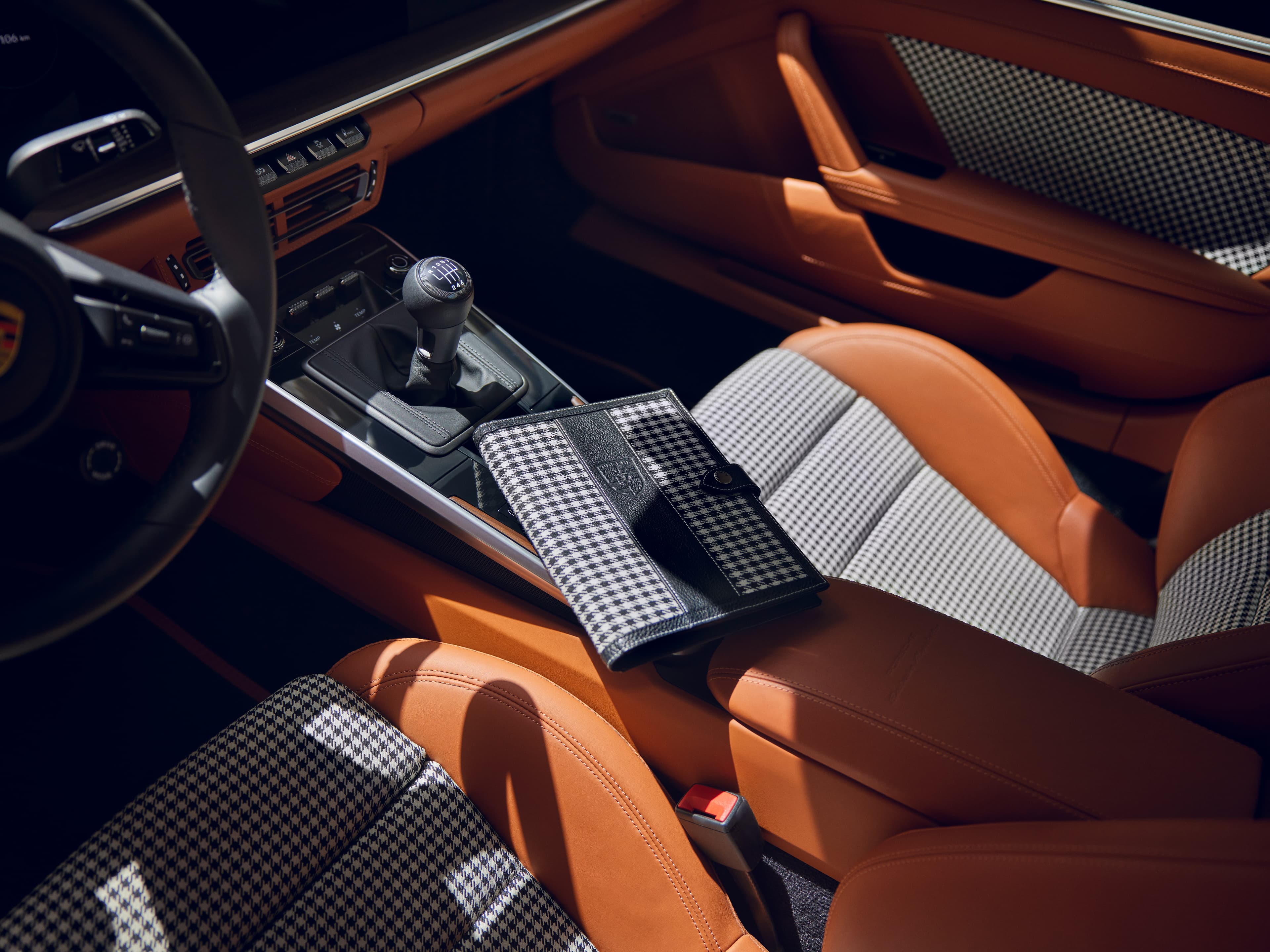 Porsche-Innenraum mit Hahnentritt-Sitzen, beigem Leder, manuellem Schalthebel und passender Hahnentritt-Clutch auf dem Beifahrersitz.