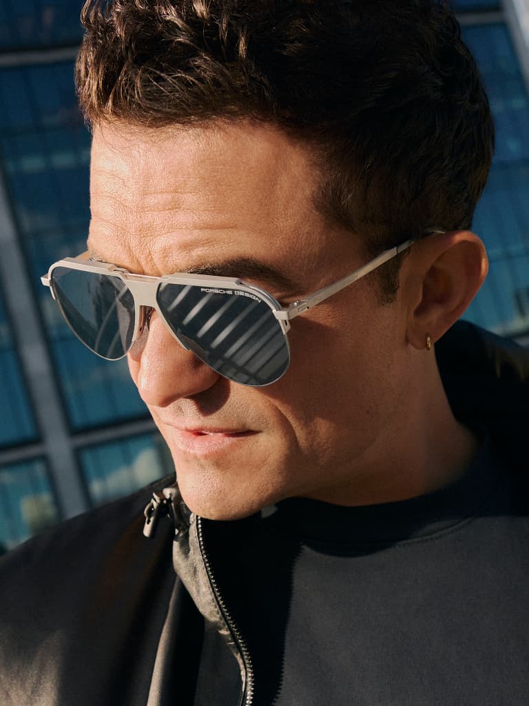 Man wearing silver Porsche Design sunglasses with dark lenses, close-up in front of a modern glass building.