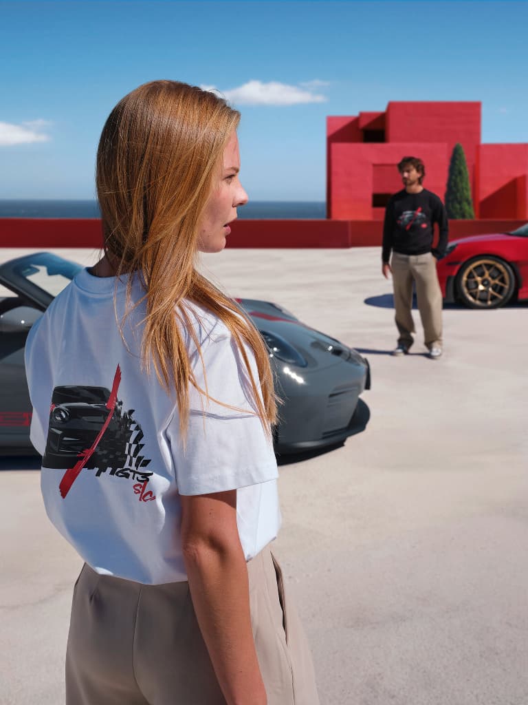 Woman wearing a white T-shirt with a back print; in the background, a man and two sports cars in front of a red seaside building.