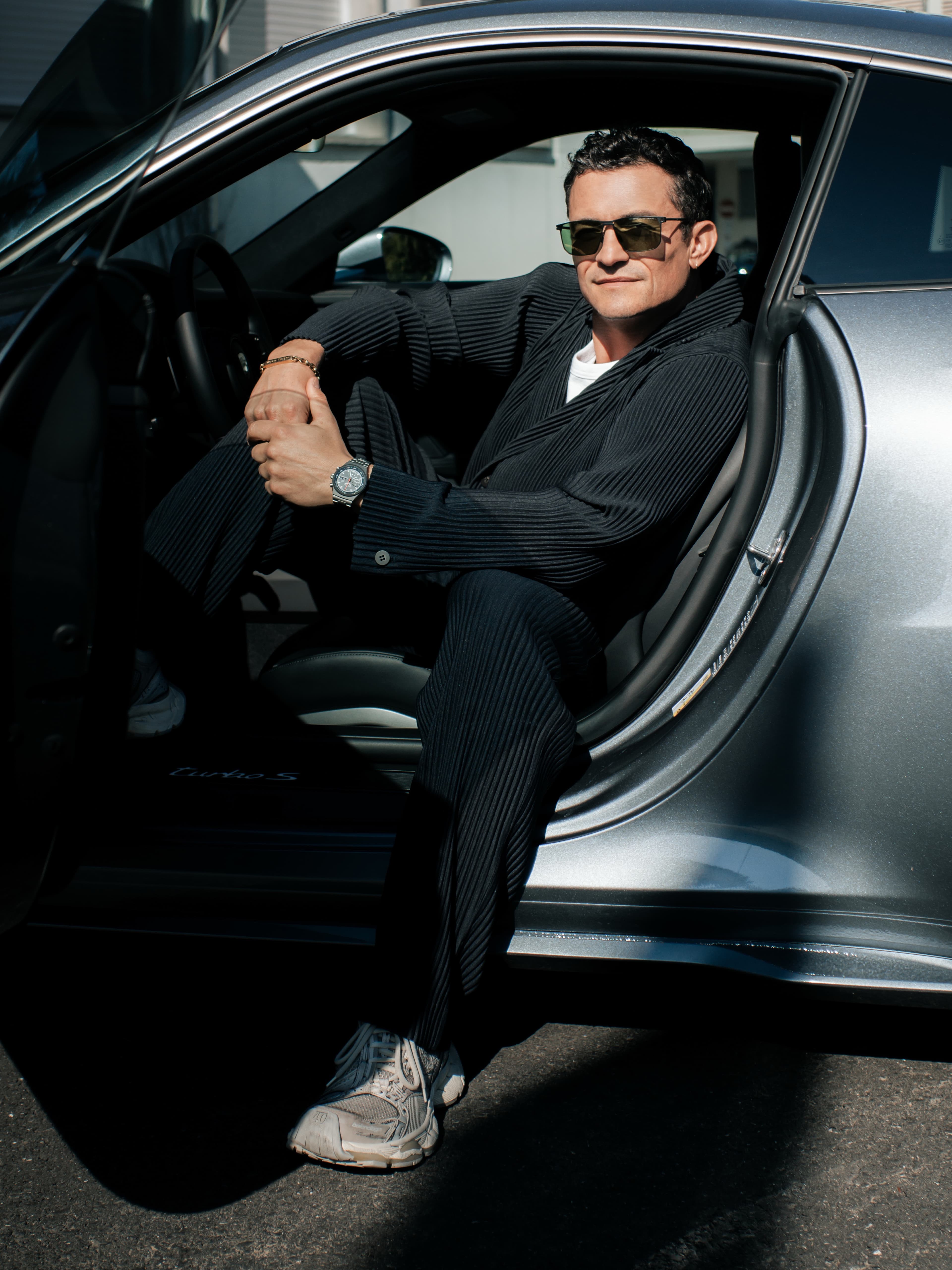 Mann mit Sonnenbrille im schwarzen Anzug sitzt in einem silbernen Porsche mit offener Tür, Armbanduhr sichtbar.