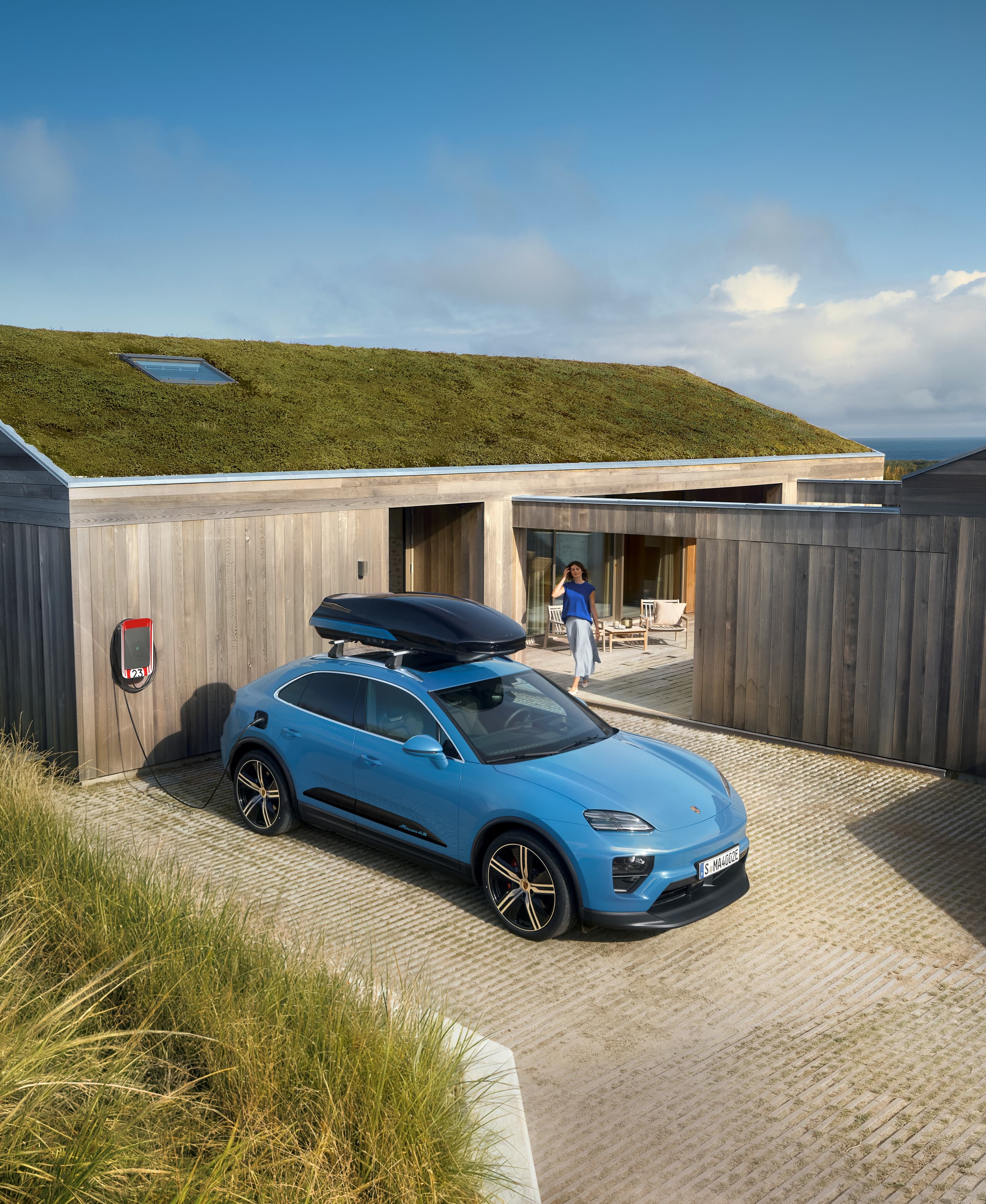 Blauwe Porsche SUV met dakbox aan een laadstation voor een modern houten huis met groen dak, persoon op het terras.