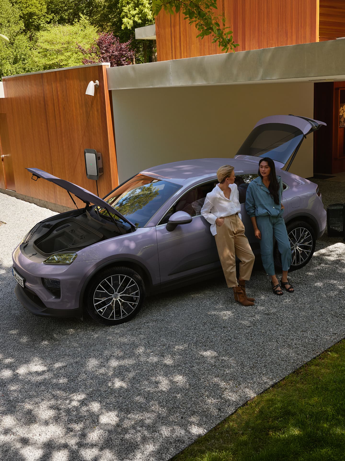 Zwei Frauen lehnen an einem lila Porsche mit offenem Kofferraum und Motorhaube, geparkt neben einem modernen Holzhaus in grüner Umgebung.