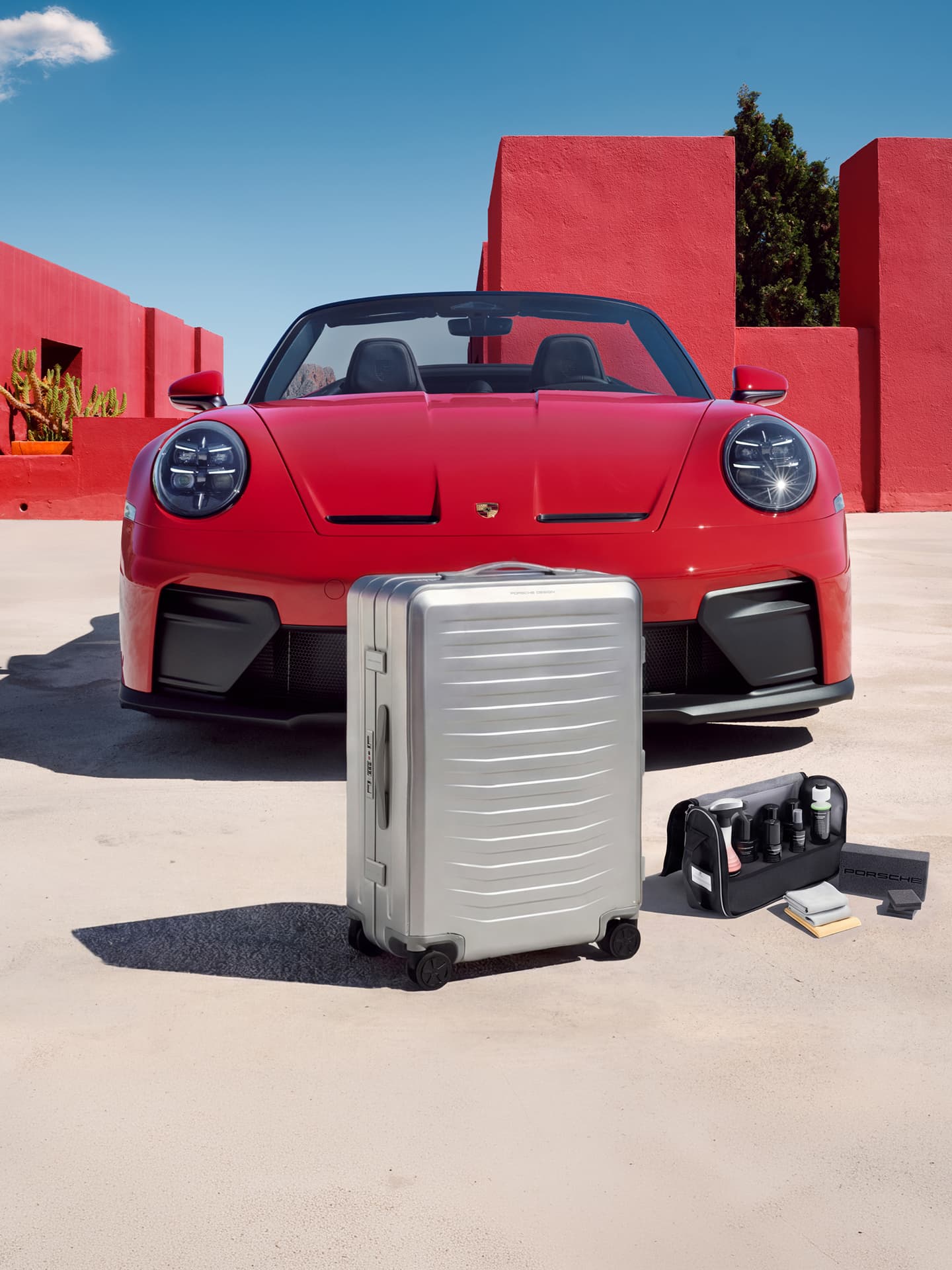 Valise rigide argentée et kit d’entretien devant un cabriolet Porsche rouge, devant des murs rouges sous un ciel bleu.