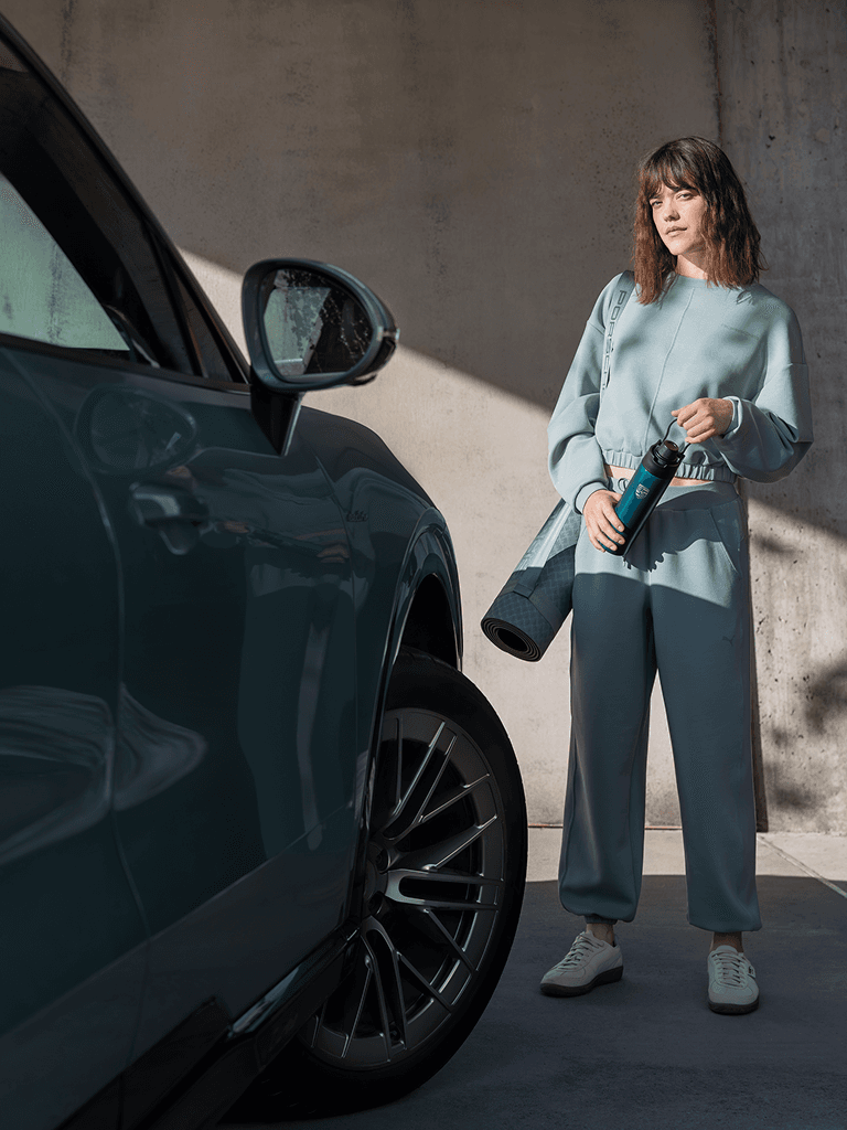Mujer con chándal azul sosteniendo una botella de agua y una esterilla de yoga, de pie junto a un coche azul bajo la luz del sol.