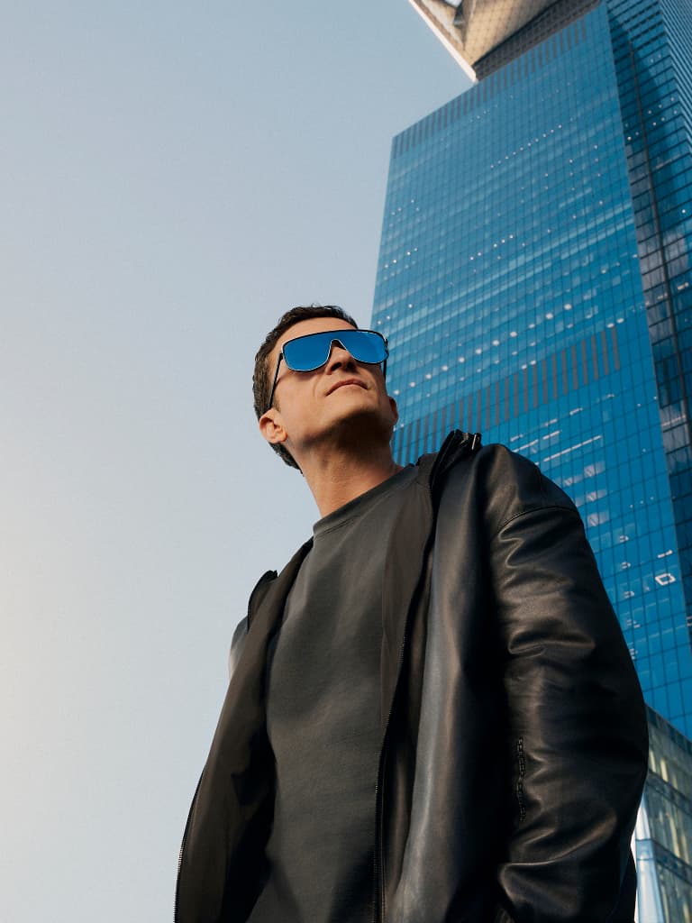 Orlando Bloom che indossa i nuovi occhiali da sole Porsche Design davanti a uno skyline cittadino