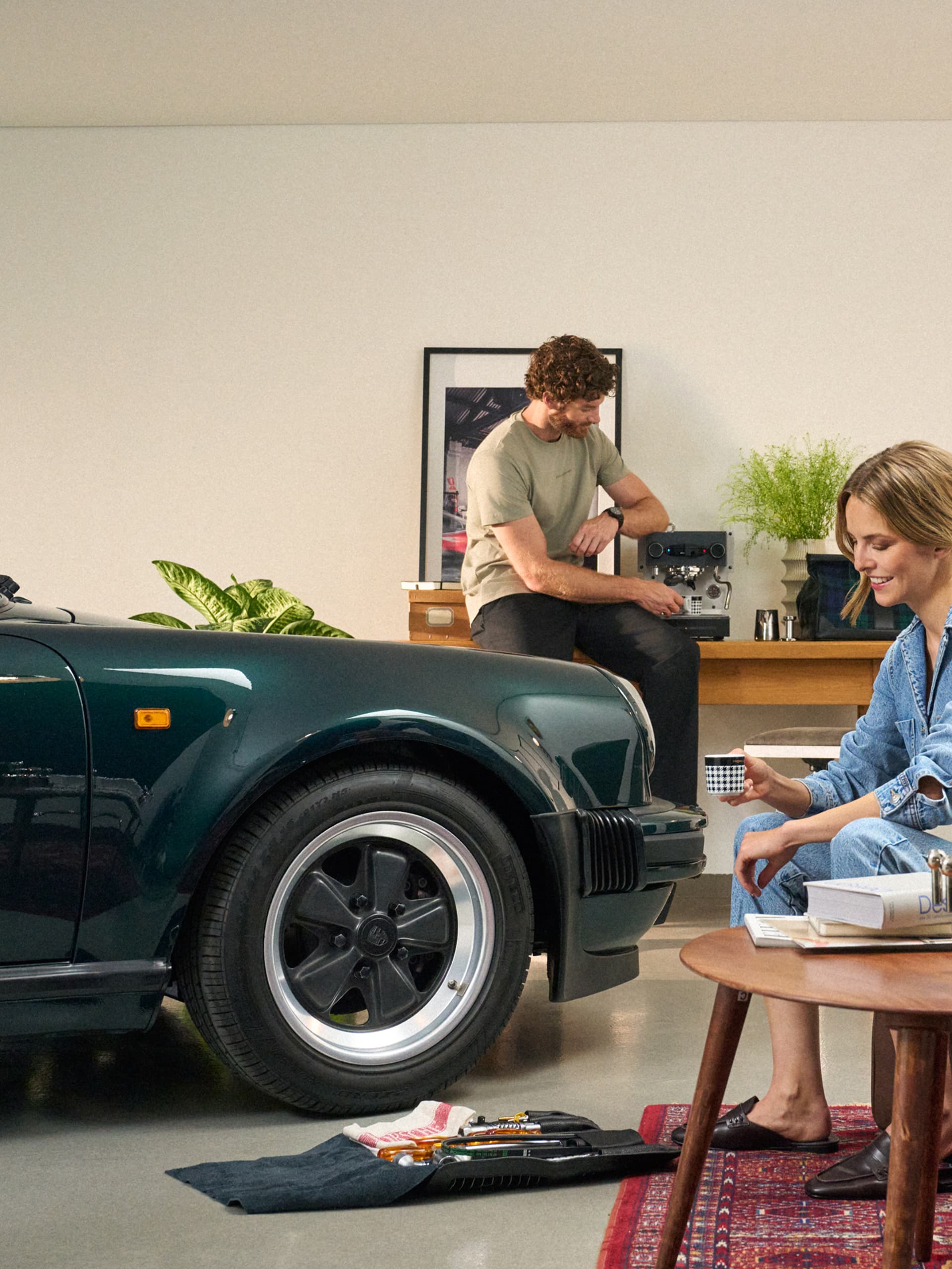Mężczyzna przygotowuje kawę przy ekspresie do espresso; kobieta siedzi z filiżanką obok ciemnozielonego Porsche w garażu.