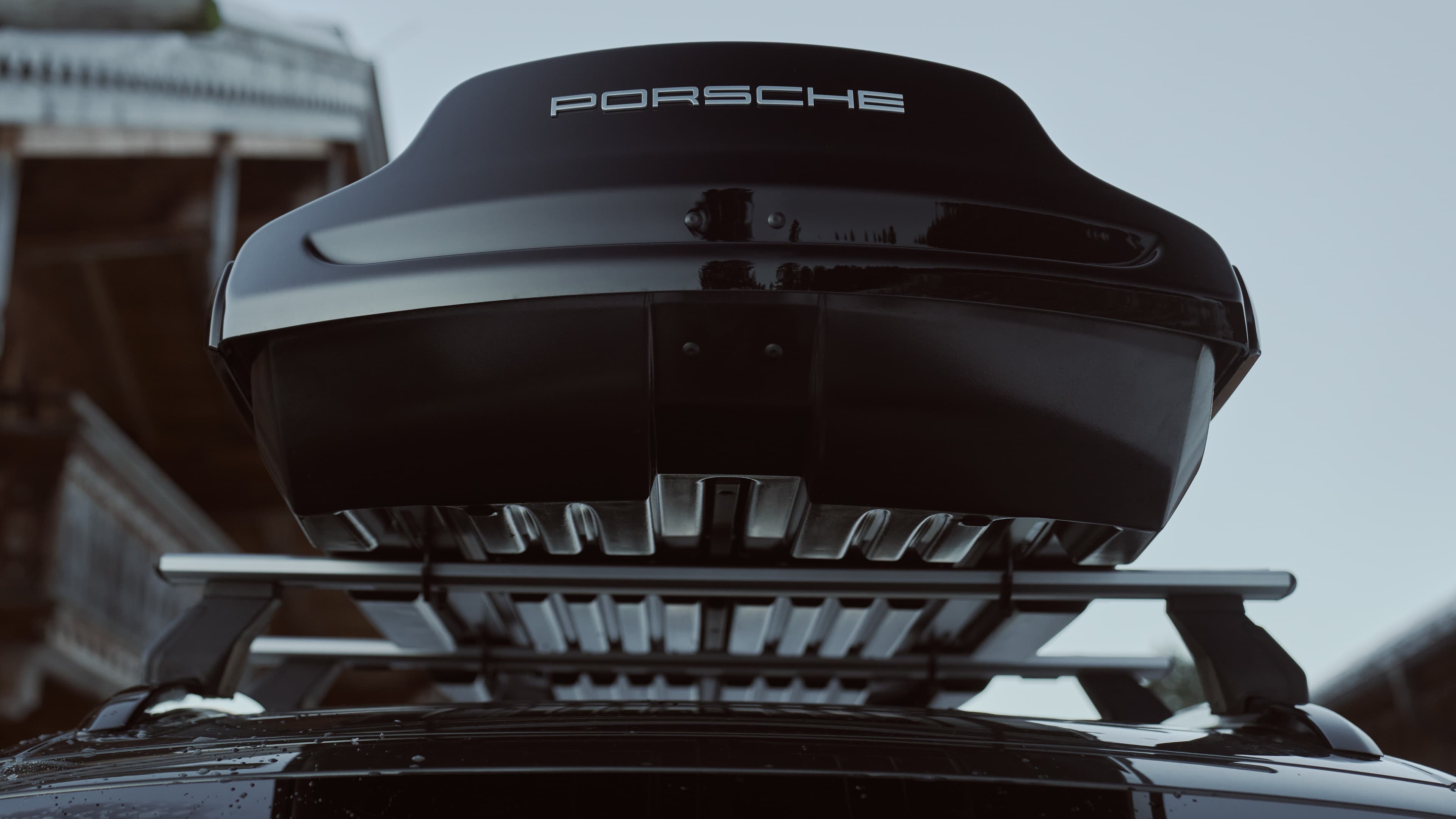 Schwarze Porsche-Dachbox auf Auto, von unten betrachtet, mit schlankem Design und sichtbarem Logo vor klarem Himmel.