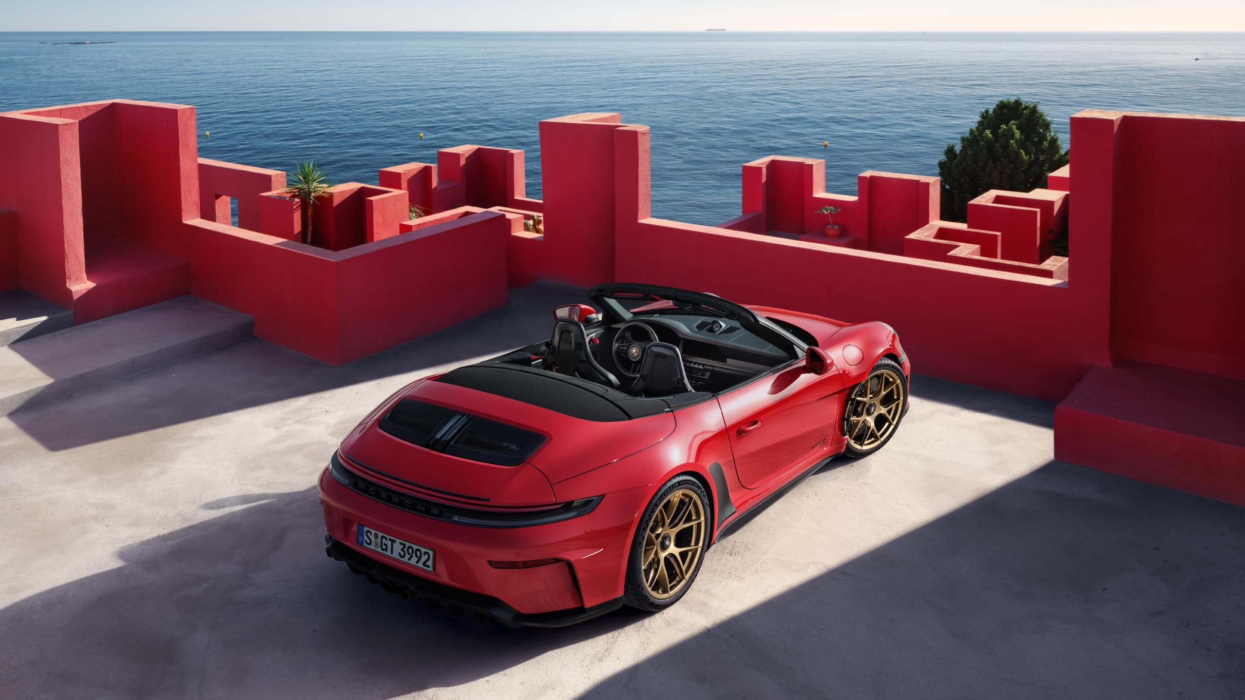 Rode Porsche 911 Cabriolet met gouden velgen, geparkeerd voor rode muren met uitzicht op zee.