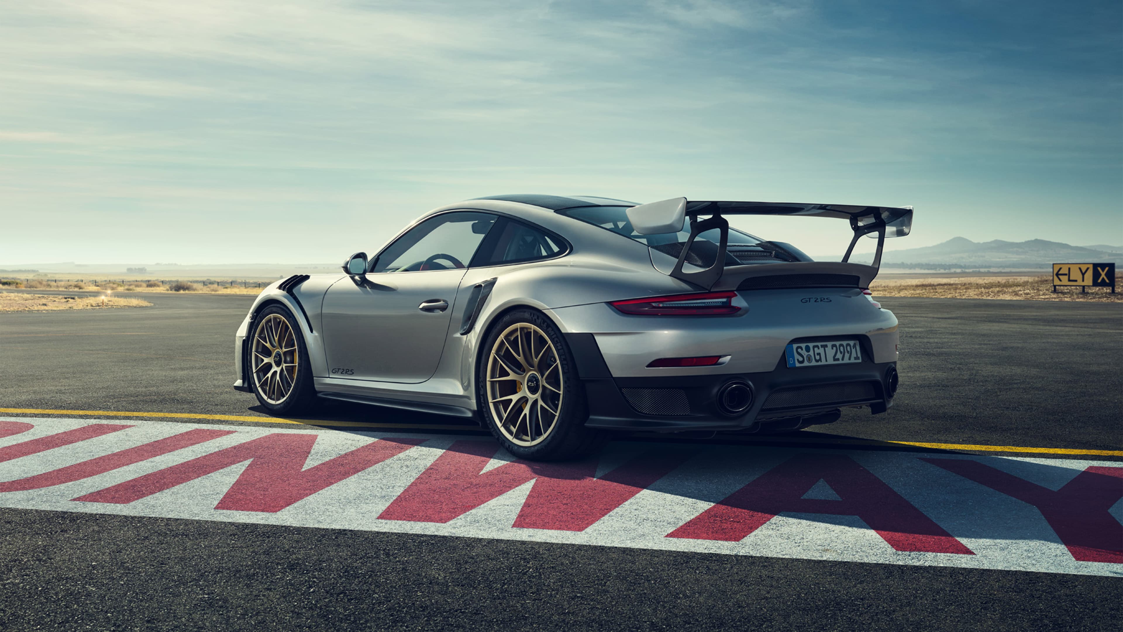 A silver Porsche 911 GT2 RS with a large rear wing on a racetrack, viewed from the rear left, with a vast sky in the background.