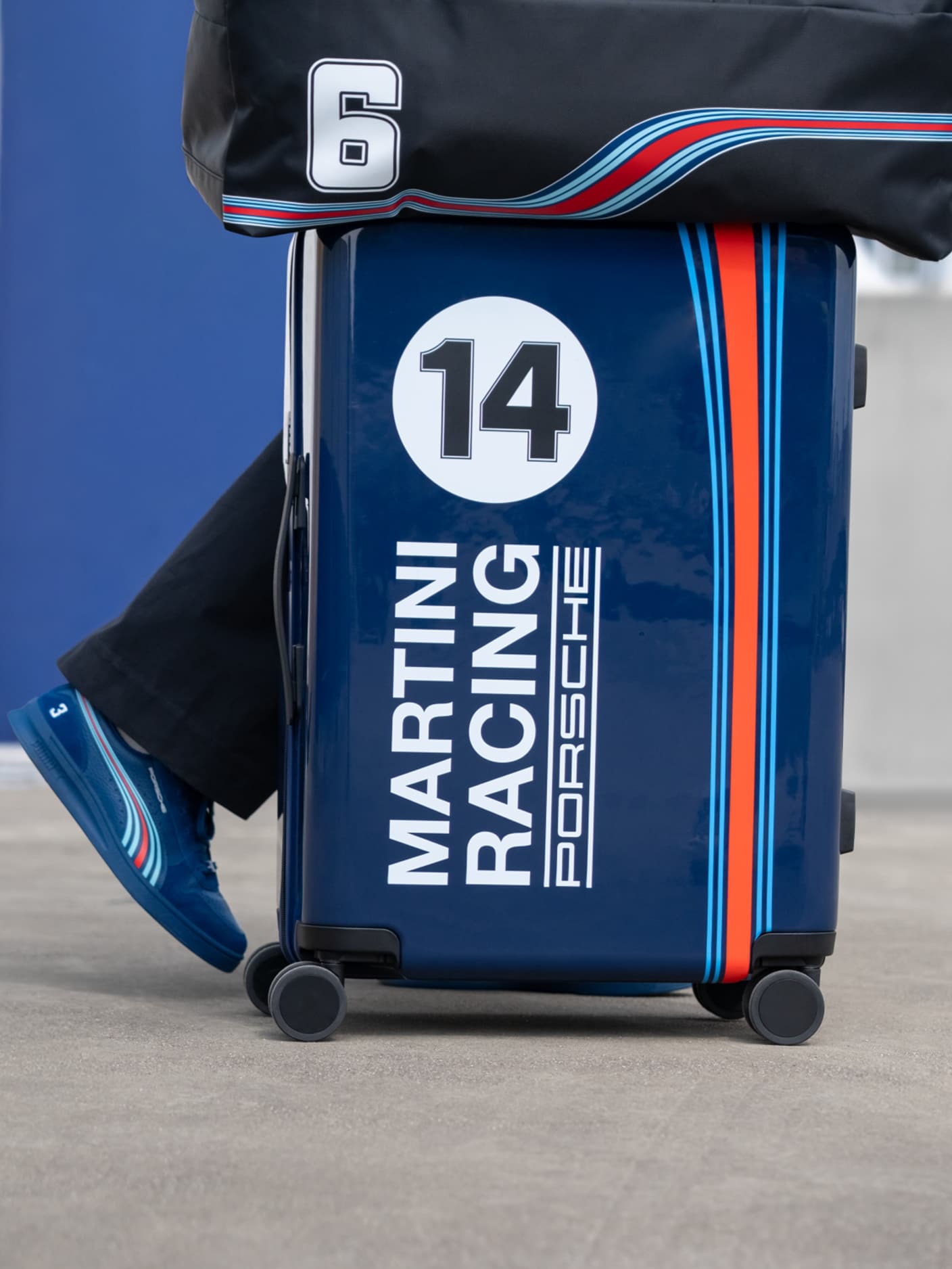 Person carring carrying a Porsche x MARTINI RACING trolley in cabin size with a duffle bag on top, wearing Porsche x MARTINI racing sneaker in blue