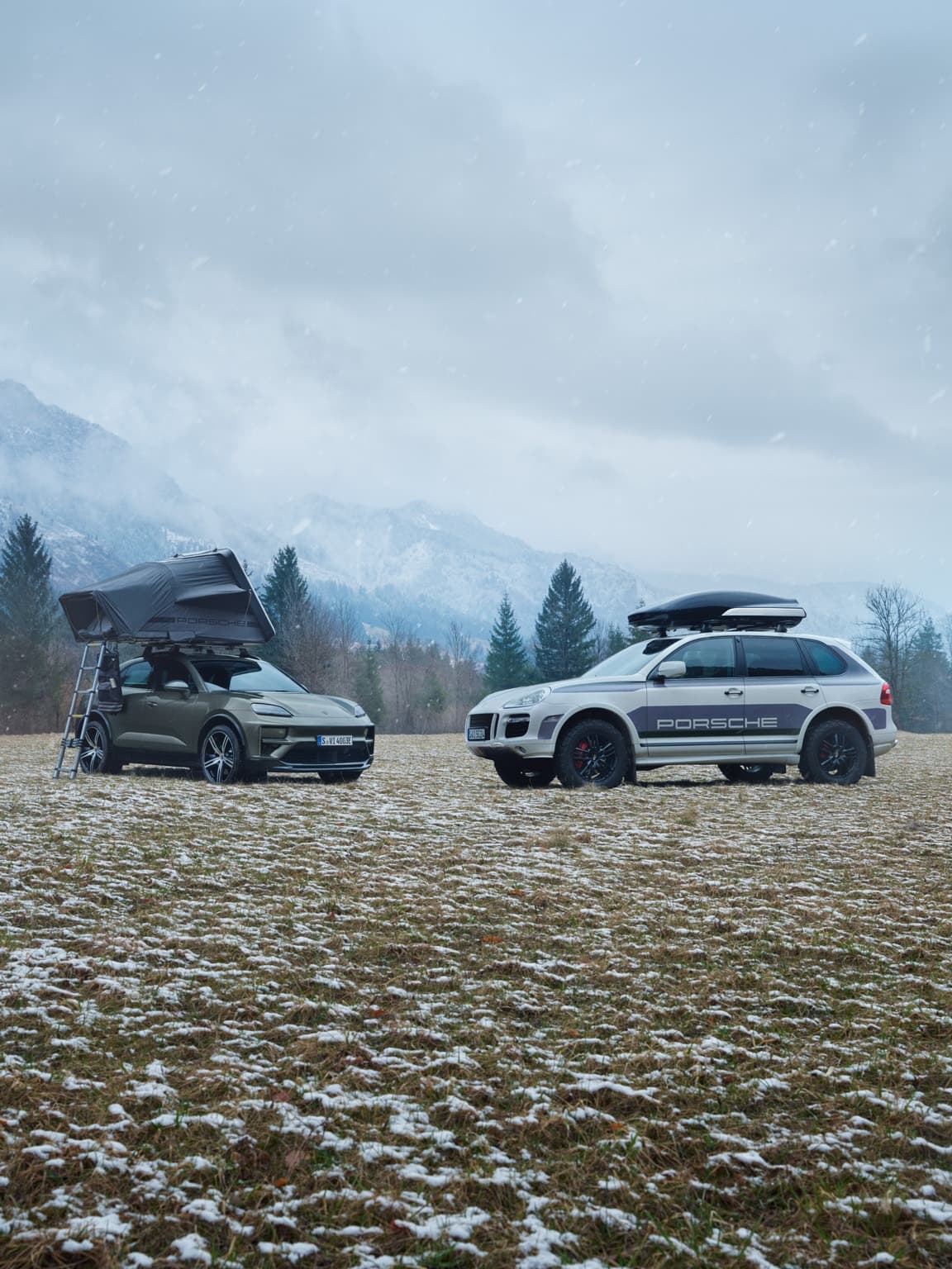 Two Porsche SUVs with roof tents in a snowy field, surrounded by mountains and trees under a cloudy sky.