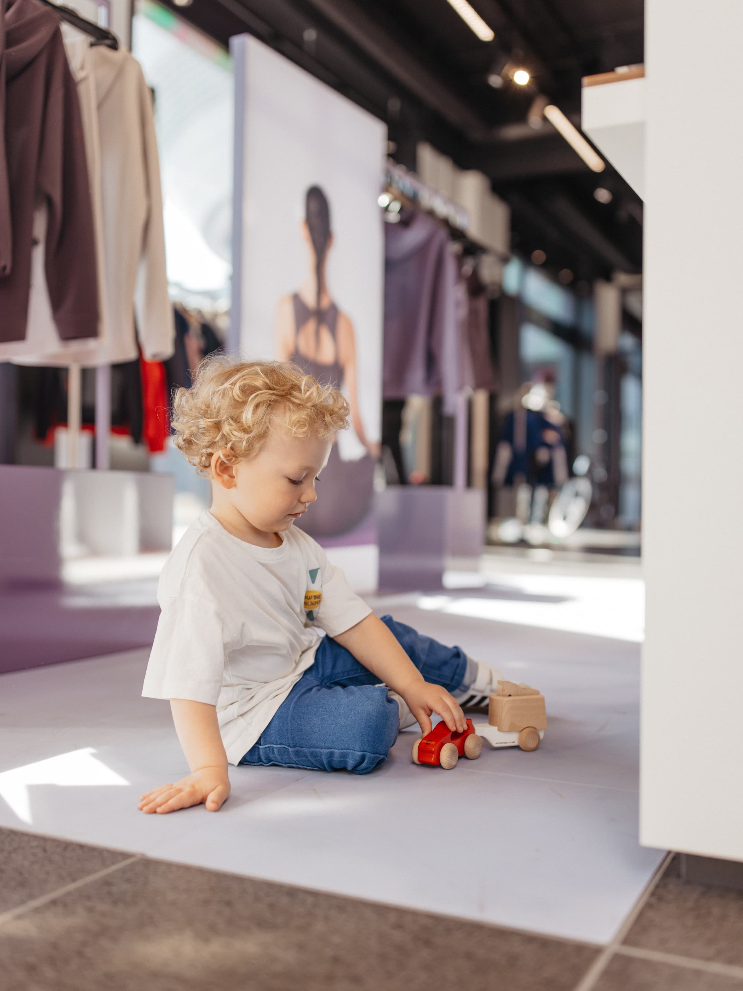 Child in white shirt and jeans plays with red toy car on store floor, surrounded by clothing displays and soft lighting.