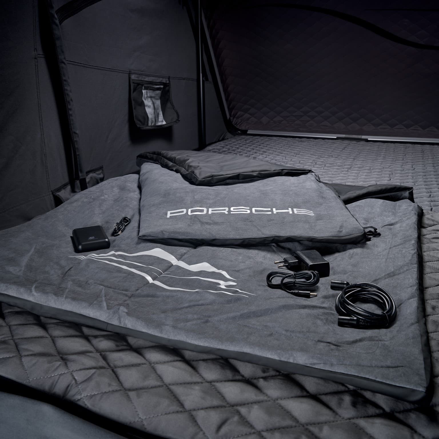Porsche-grauer Schlafsack mit Ladegerät und Kabeln auf gestepptem Untergrund in einem dunklen Zelt.