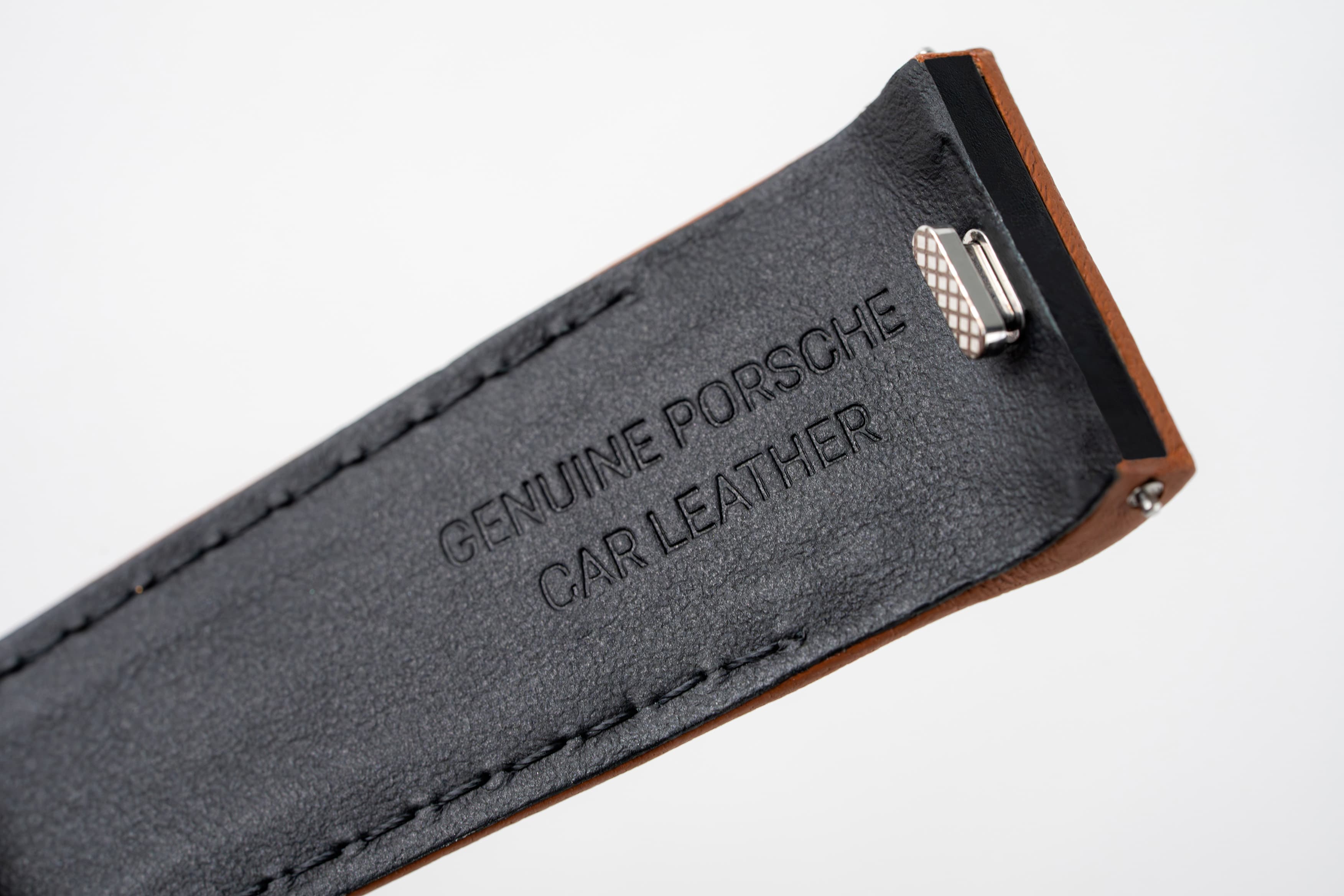 Close-up van een zwarte leren band met reliëftekst “Genuine Porsche Car Leather” en een metalen clip.