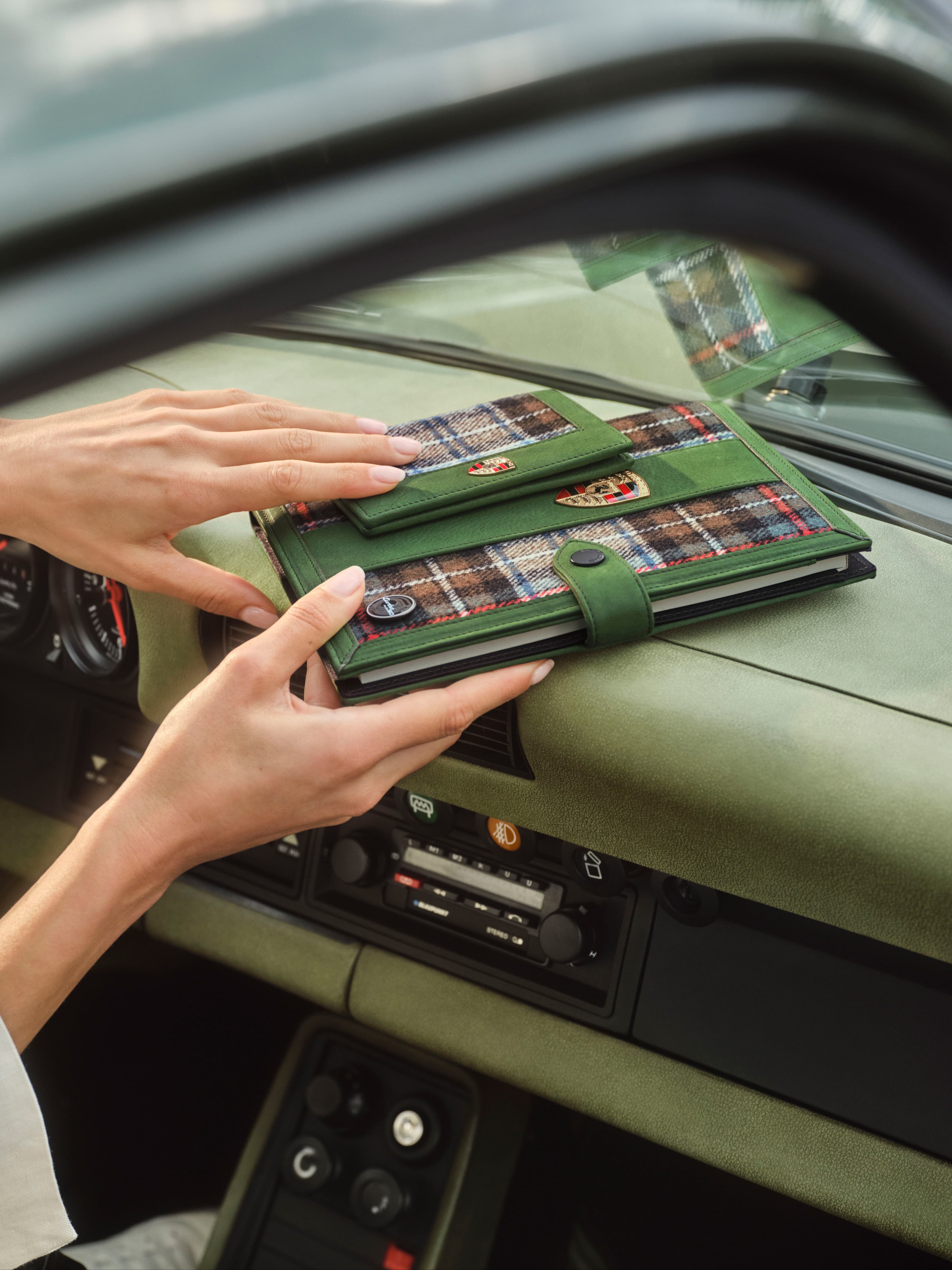 Hands holding green leather and tartan notebook cover with Porsche crest inside a car dashboard view