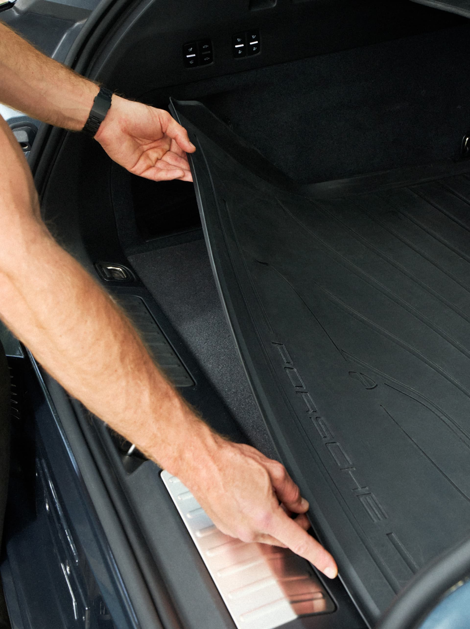 Mano che posiziona un rivestimento bagagliaio nero Porsche nel vano bagagli posteriore, primo piano del tappetino in gomma testurizzata e della soglia di carico.