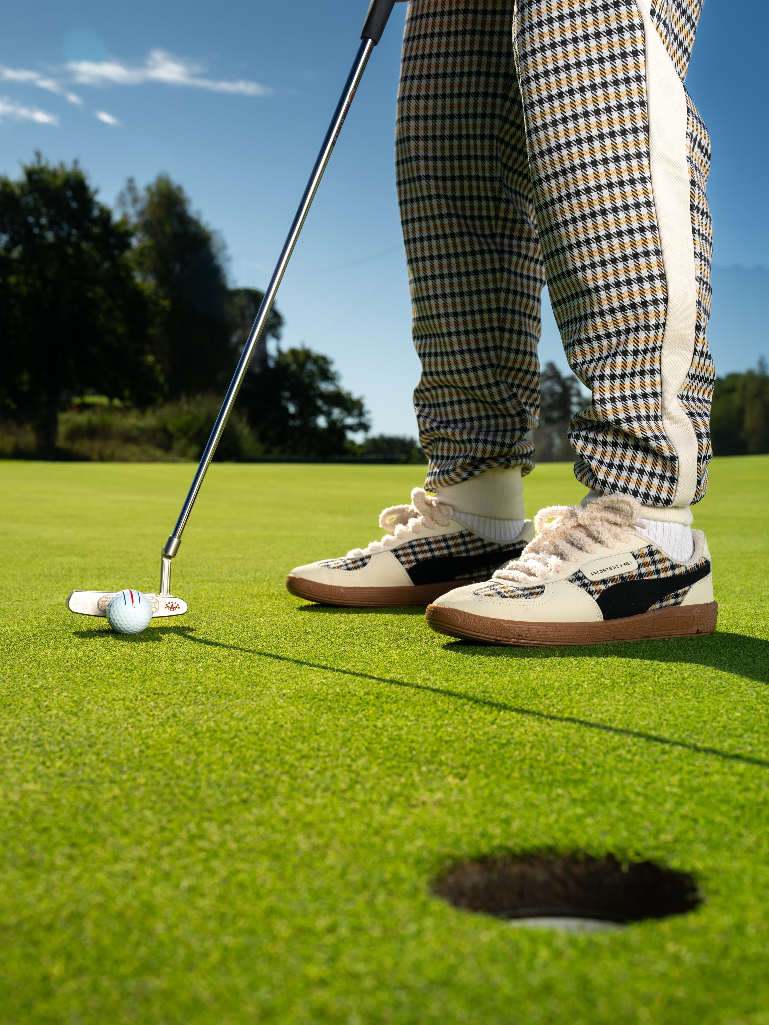 Person in karierten Hosen und Sneakers, die einen Golfball auf einem grünen Platz unter klarem blauen Himmel puttet.
