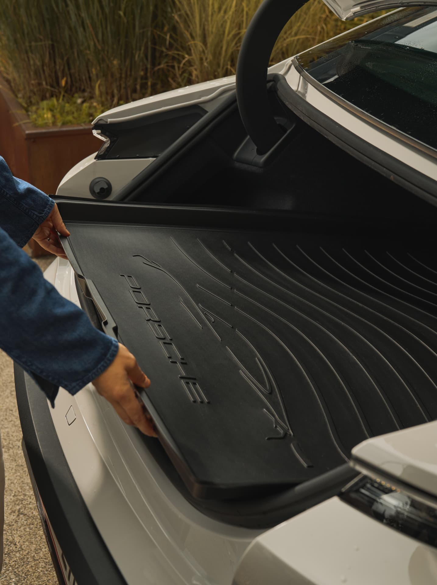 Person placing a black Porsche trunk liner in a white car, with greenery in the background.