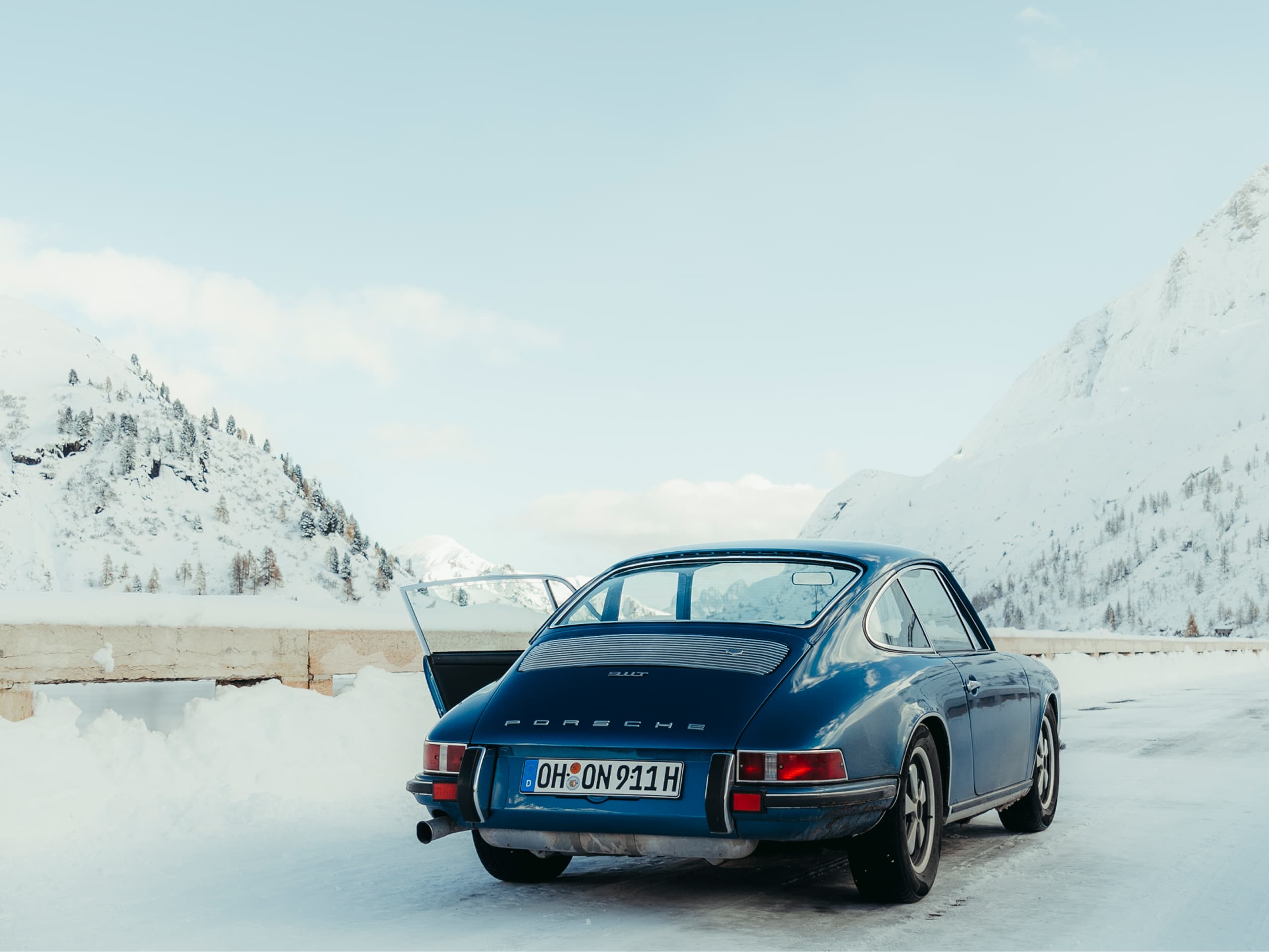 Blue Porsche 911 parked on snowy mountain road, open door, clear sky, snow-covered peaks in background.