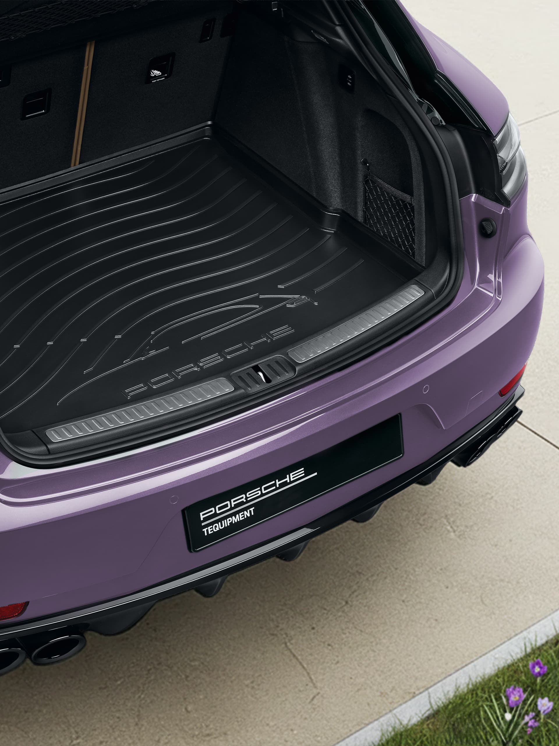 Coffre ouvert d'une Porsche violette avec doublure de coffre noire, sur une allée en pierre claire avec de l'herbe et des fleurs violettes à proximité.