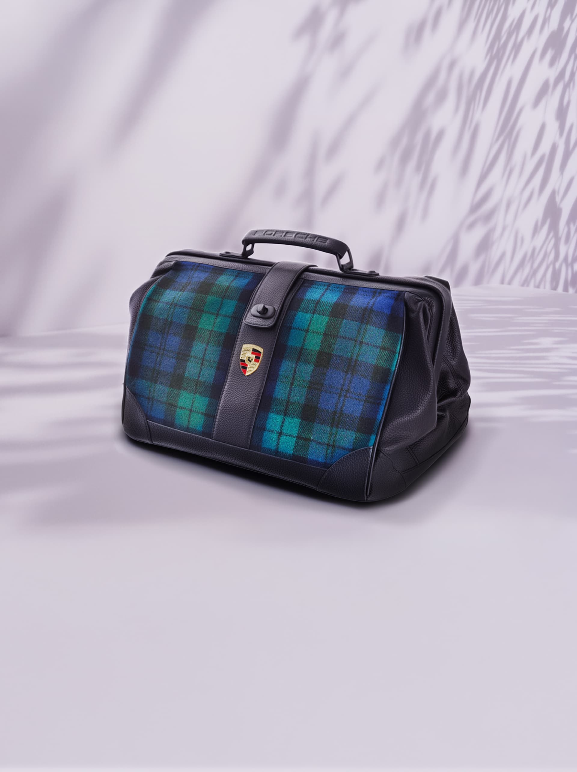 Zwarte lederen tas met blauw-groen geruit patroon, Porsche-logo, op een lichte achtergrond met schaduwpatronen.