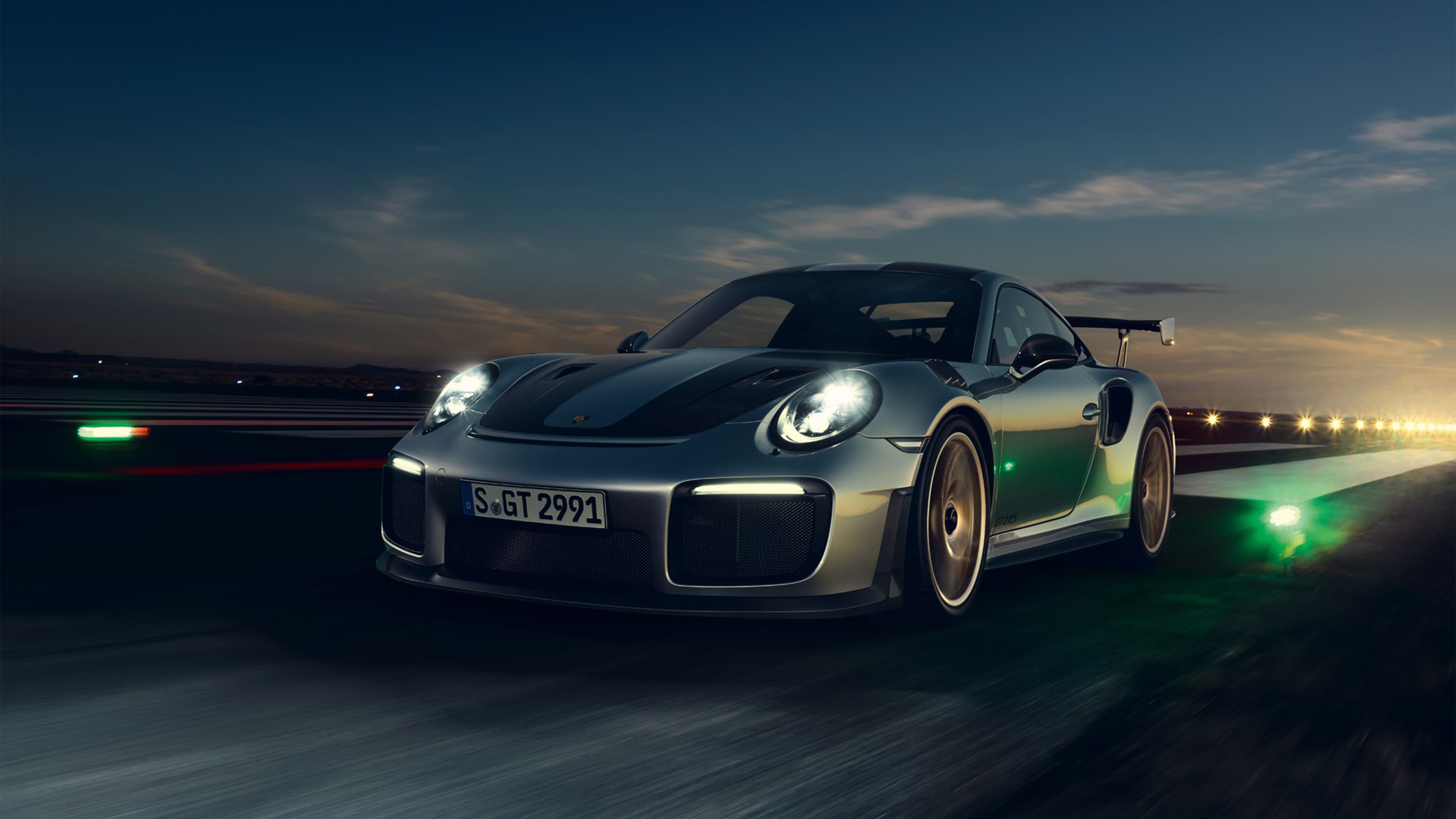 A silver Porsche 911 GT2 RS drives around the racetrack at dusk, headlights on, motion blur.