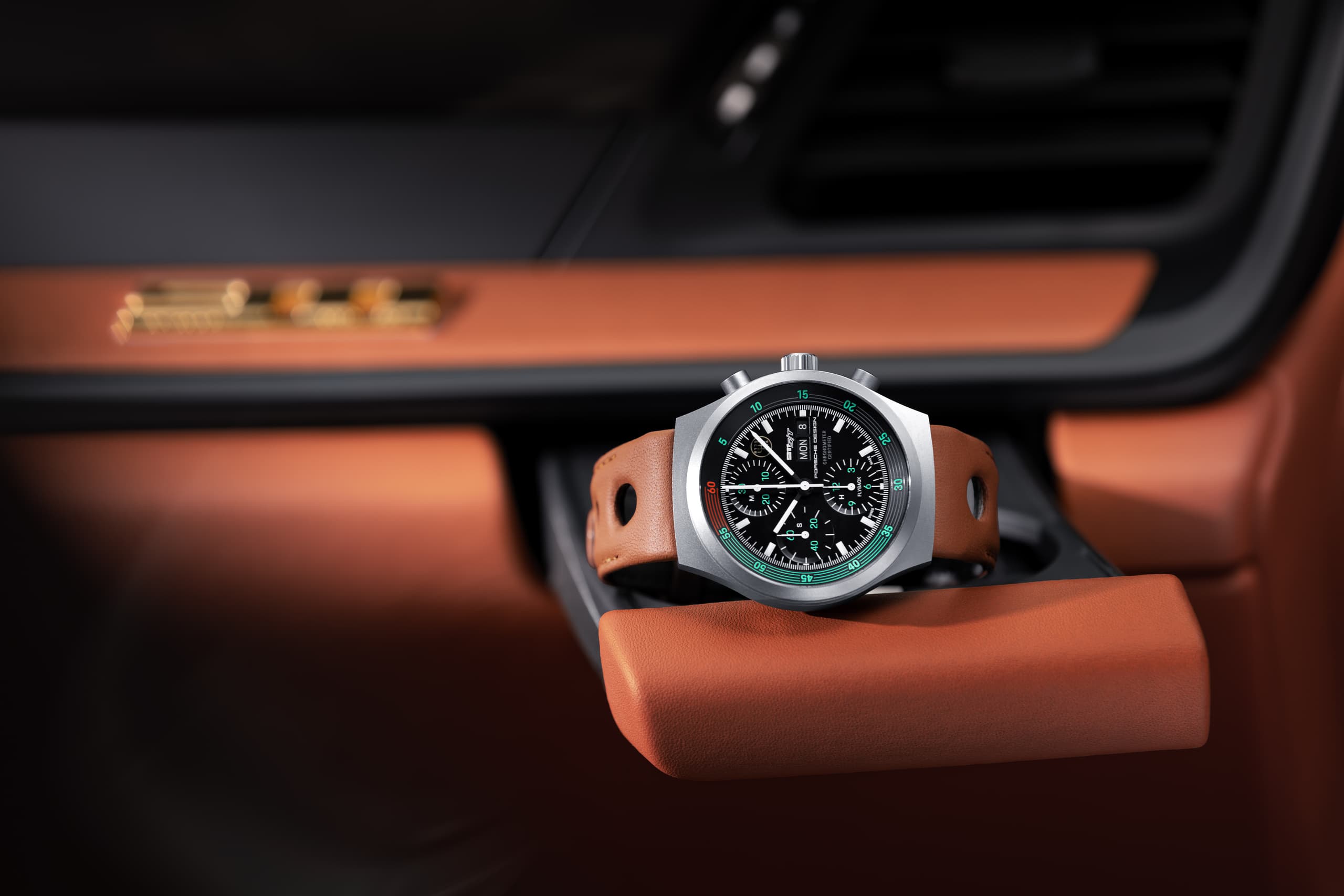 Zegarek chronograf z czarną tarczą i brązowym skórzanym paskiem, leżący na podłokietniku we wnętrzu samochodu Porsche w kolorze jasnobrązowym.