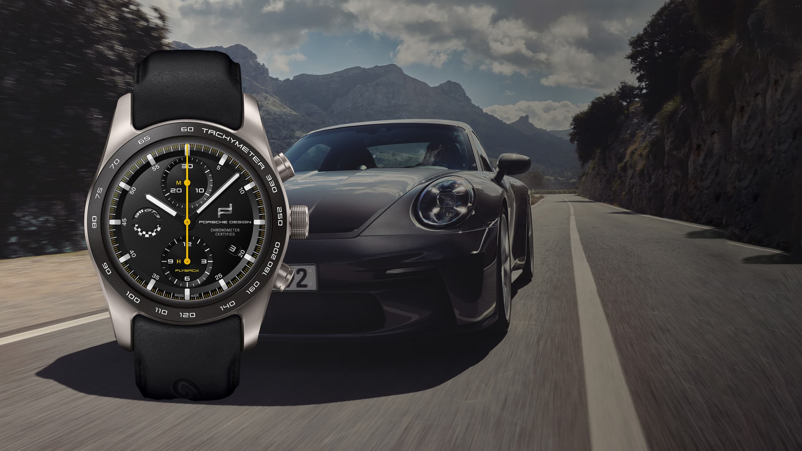 Chronograf Porsche Design z czarnym paskiem i pierścieniem tachymetru, na tle szarego sportowego samochodu na górskiej drodze