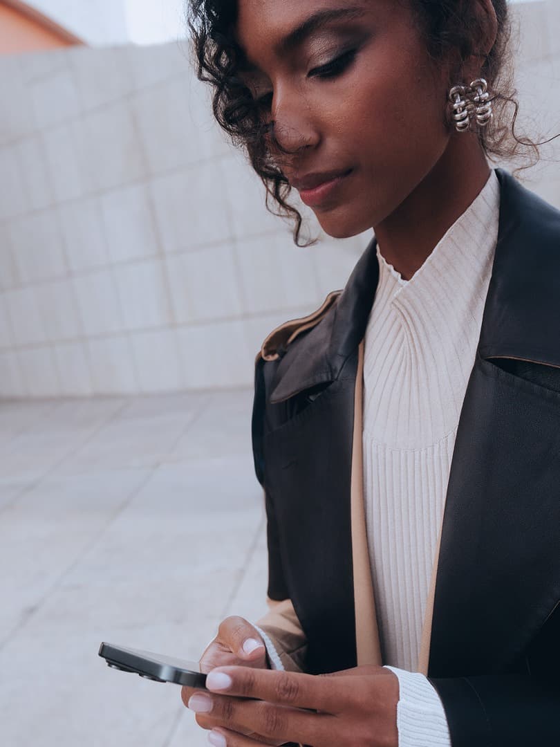 Woman in a black leather jacket and white top looking at a smartphone standing against a tiled wall background