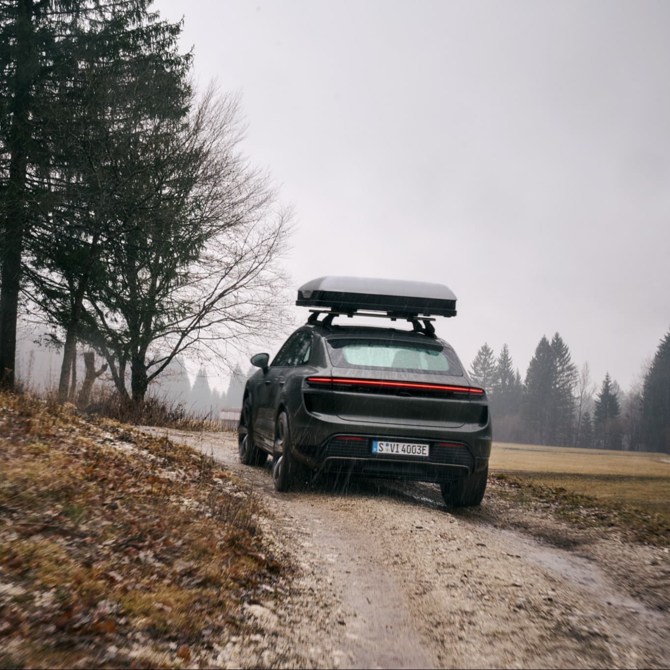 Porsche car with roof box driving on a muddy path, surrounded by trees on a cloudy day
