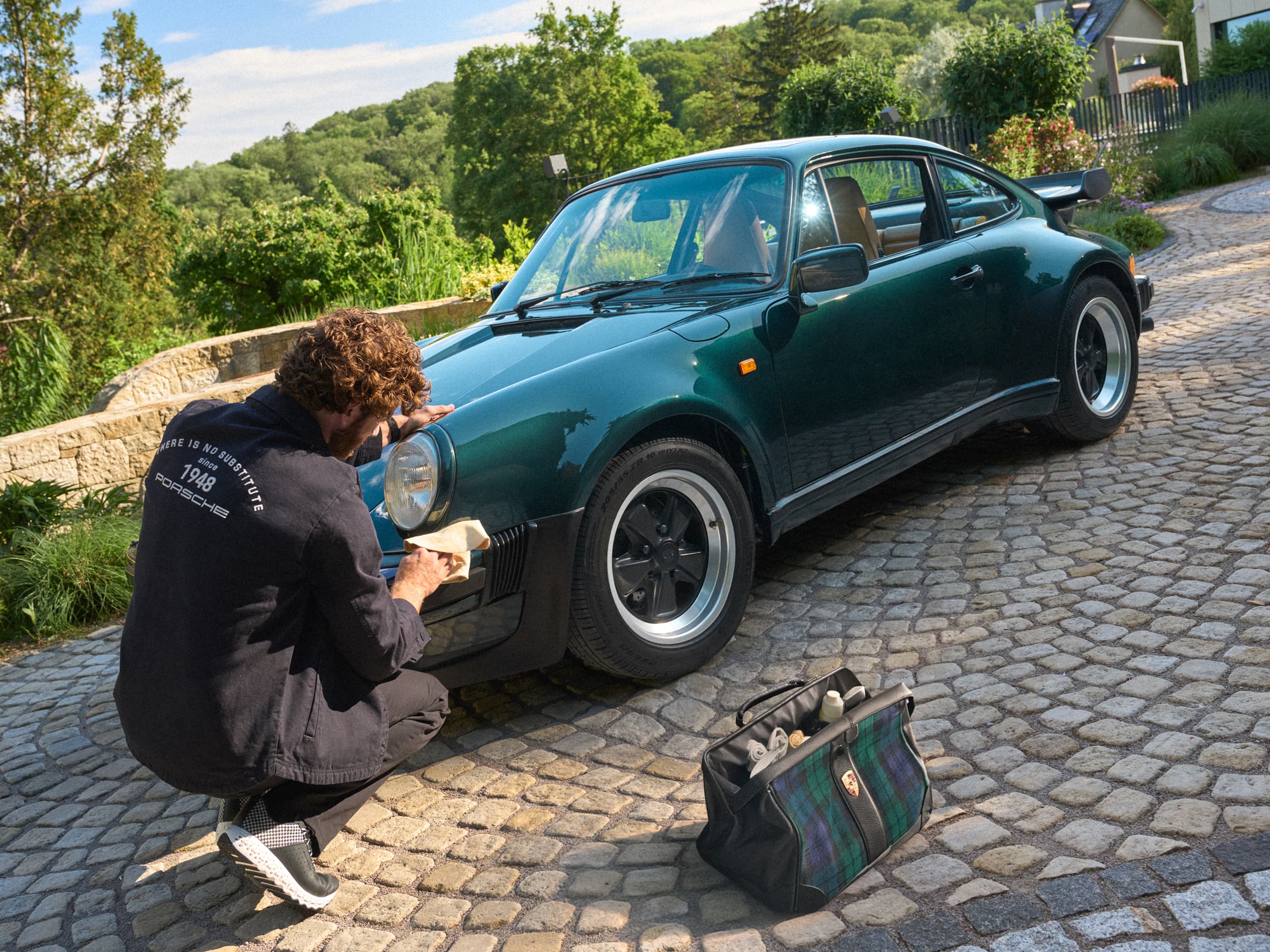 Person polishing a dark green classic Porsche 911 on a cobblestone driveway, with a tartan Porsche bag nearby