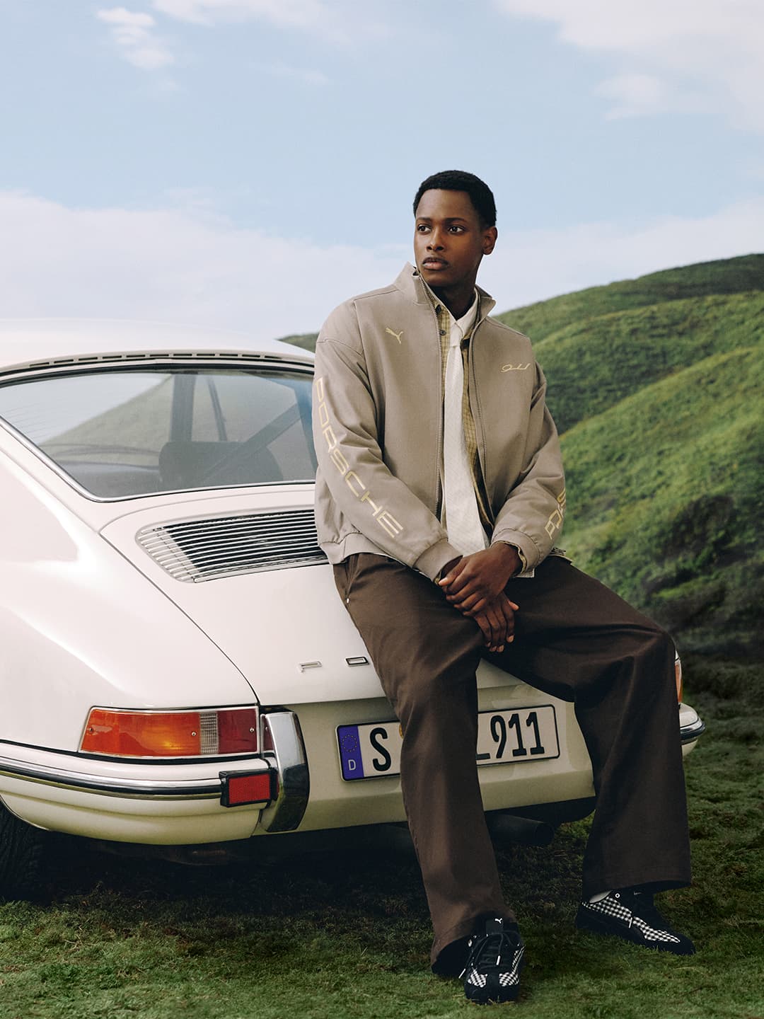 Model in beige Porsche jacket and brown pants sits on white Porsche 911 rear, green hills and blue sky behind.