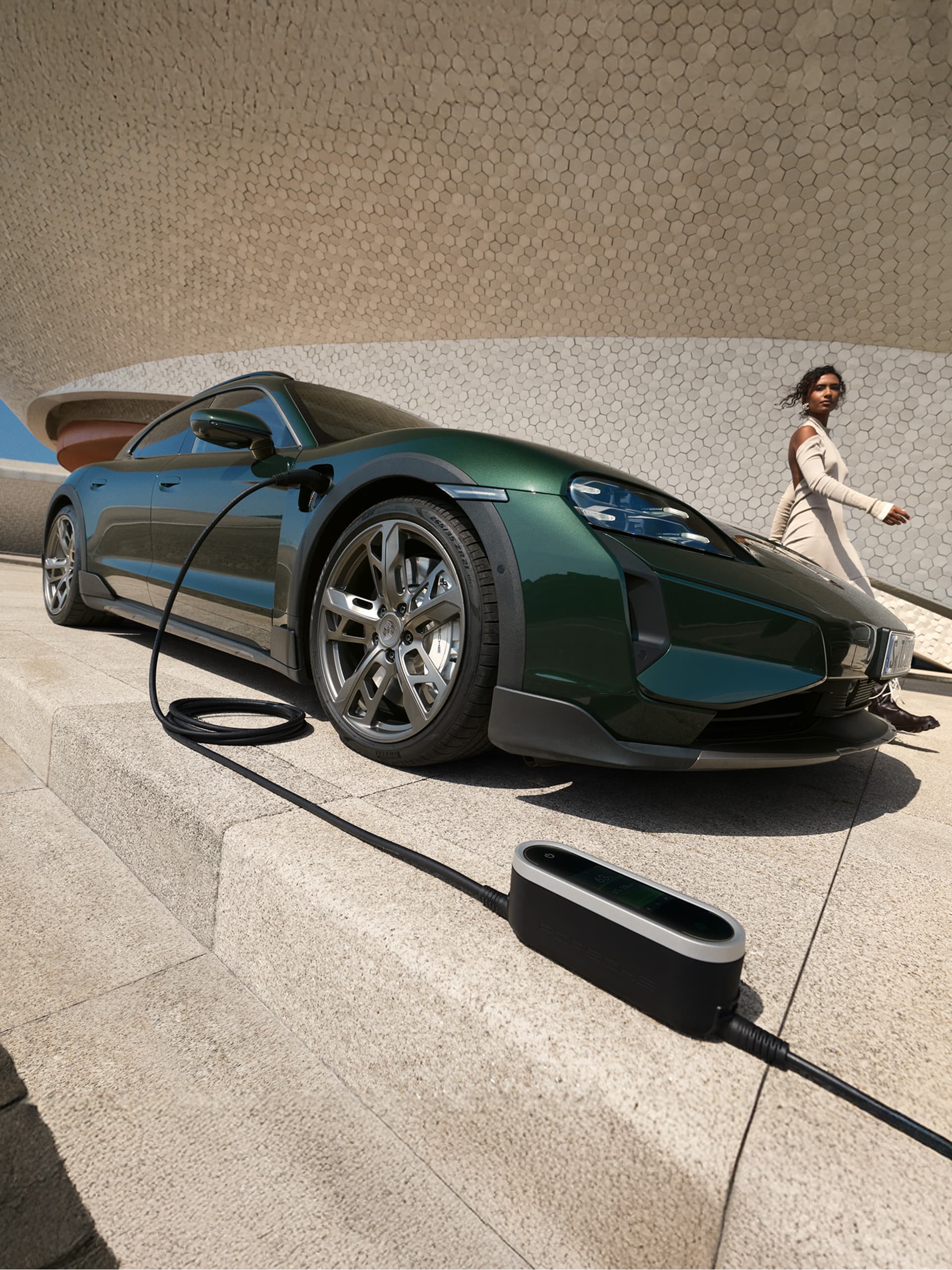 You can see a darkgreen Porsche Taycan with a Porsche Mobile Charger Connect on the ground, in front of a building