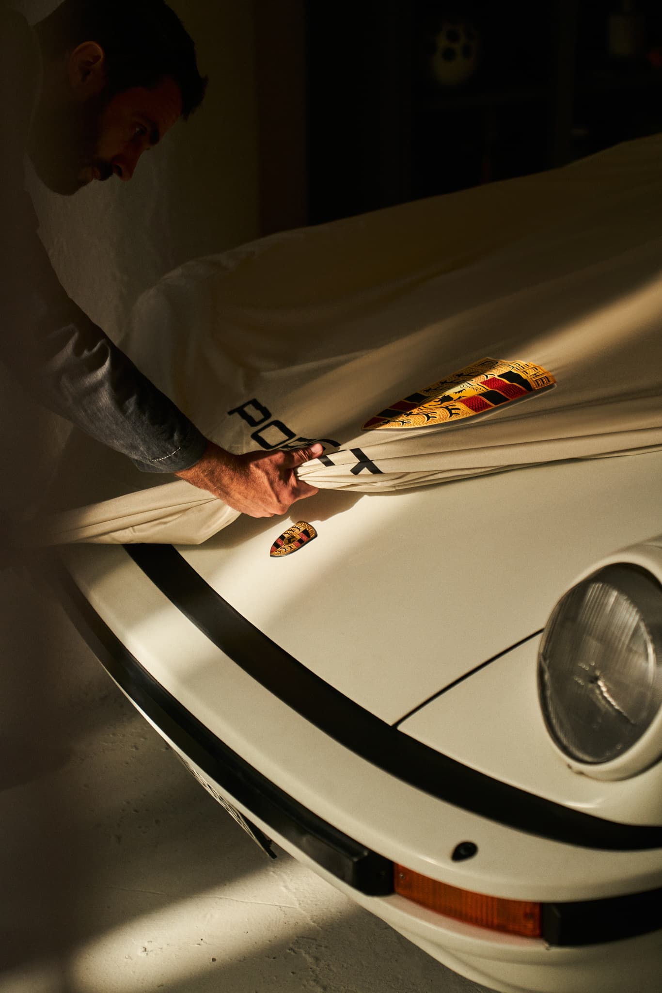 Uomo che rimuove un copriauto beige Porsche da una Porsche classica bianca con striscia nera, stemma visibile sul cofano.