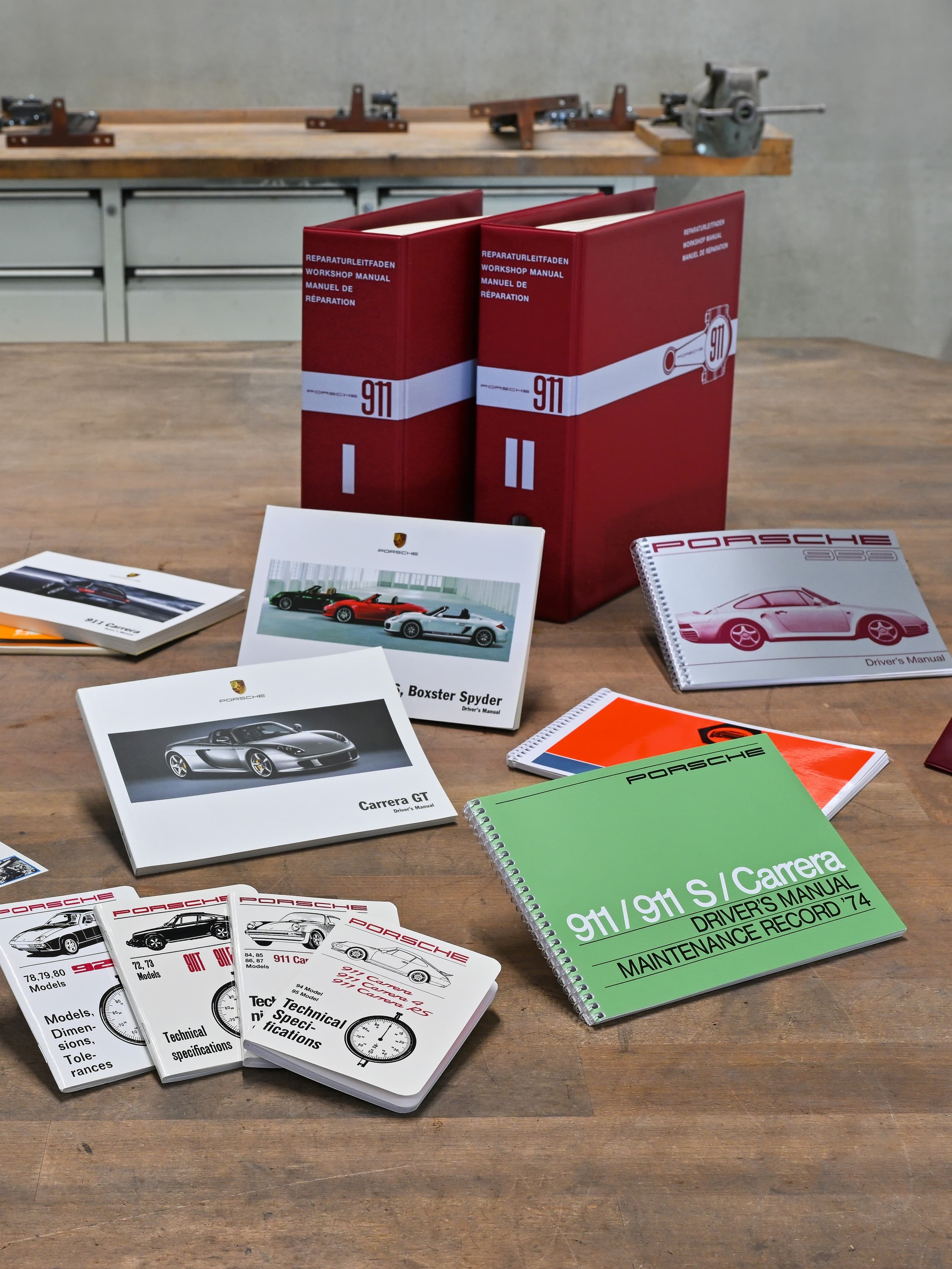 Manuels et brochures Porsche sur une table en bois avec des classeurs rouges, images de voitures et dossiers d'entretien dans un atelier.