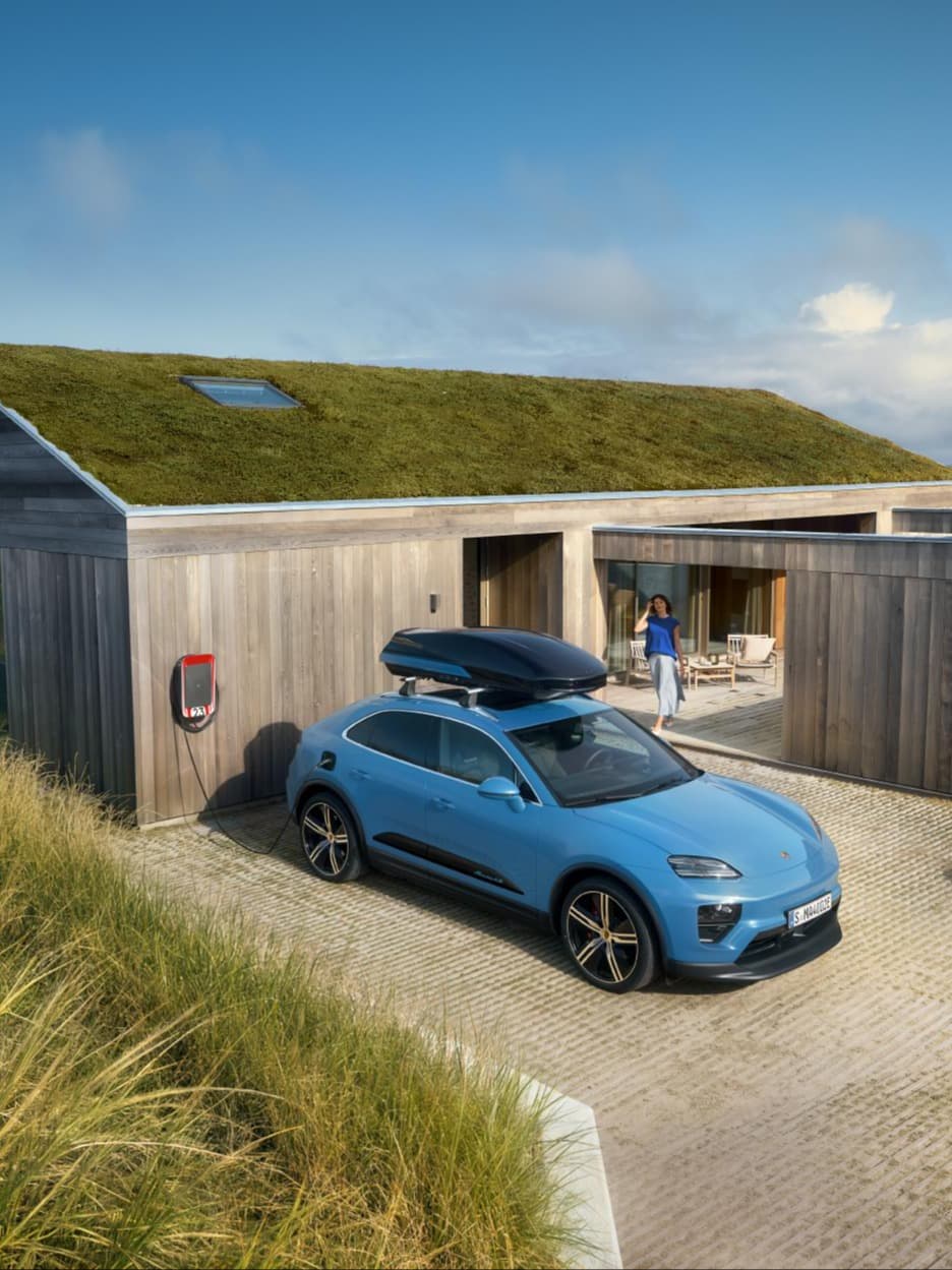 Blauwe Porsche SUV met dakbox laadt aan een wallbox bij een houten huis met groen dak; vrouw staat op het terras.