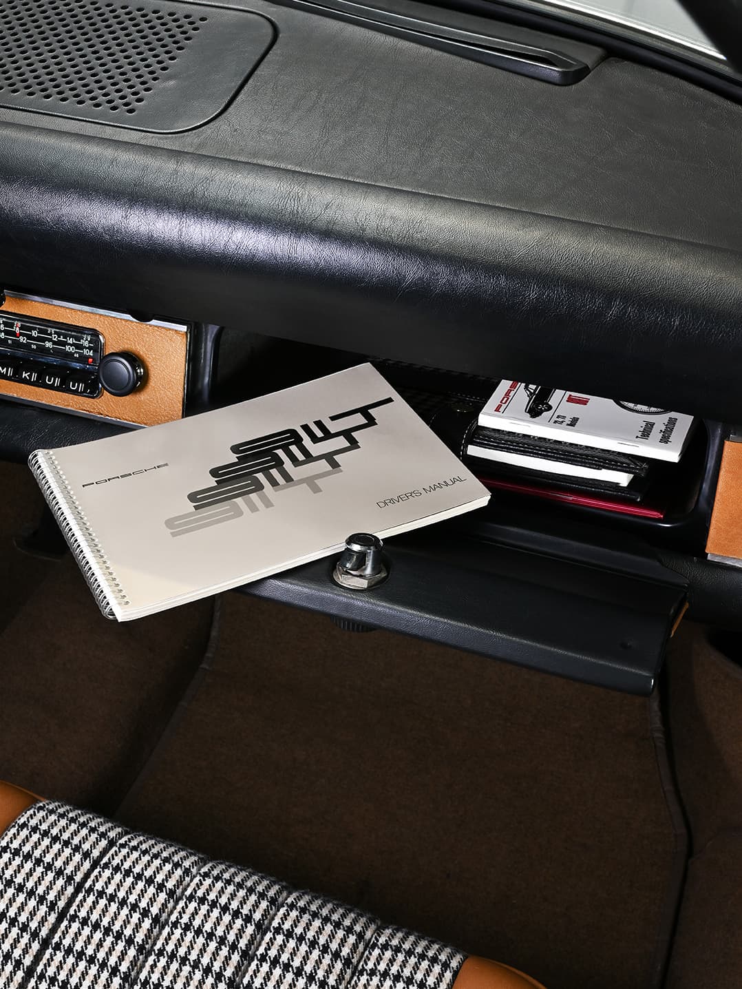 Boîte à gants ouverte avec manuel Porsche et documents à l'intérieur, tableau de bord en cuir noir, radio vintage, siège à motif pied-de-poule.
