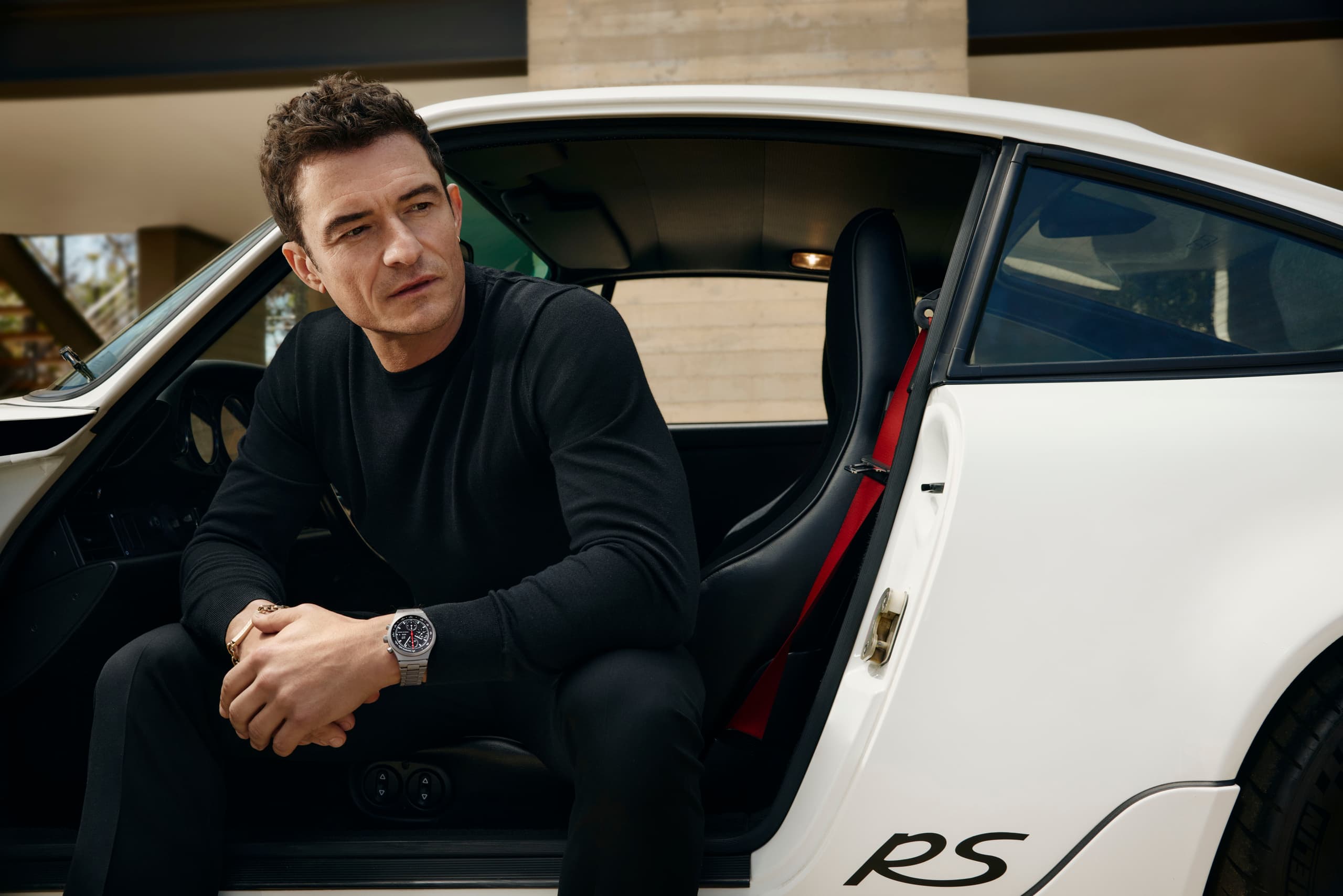 Orlando Bloom sitting in the open door of his Porsche 911 wearing the Porsche Design Chronograph 1 - 1975 Limited Edition on his wrist.