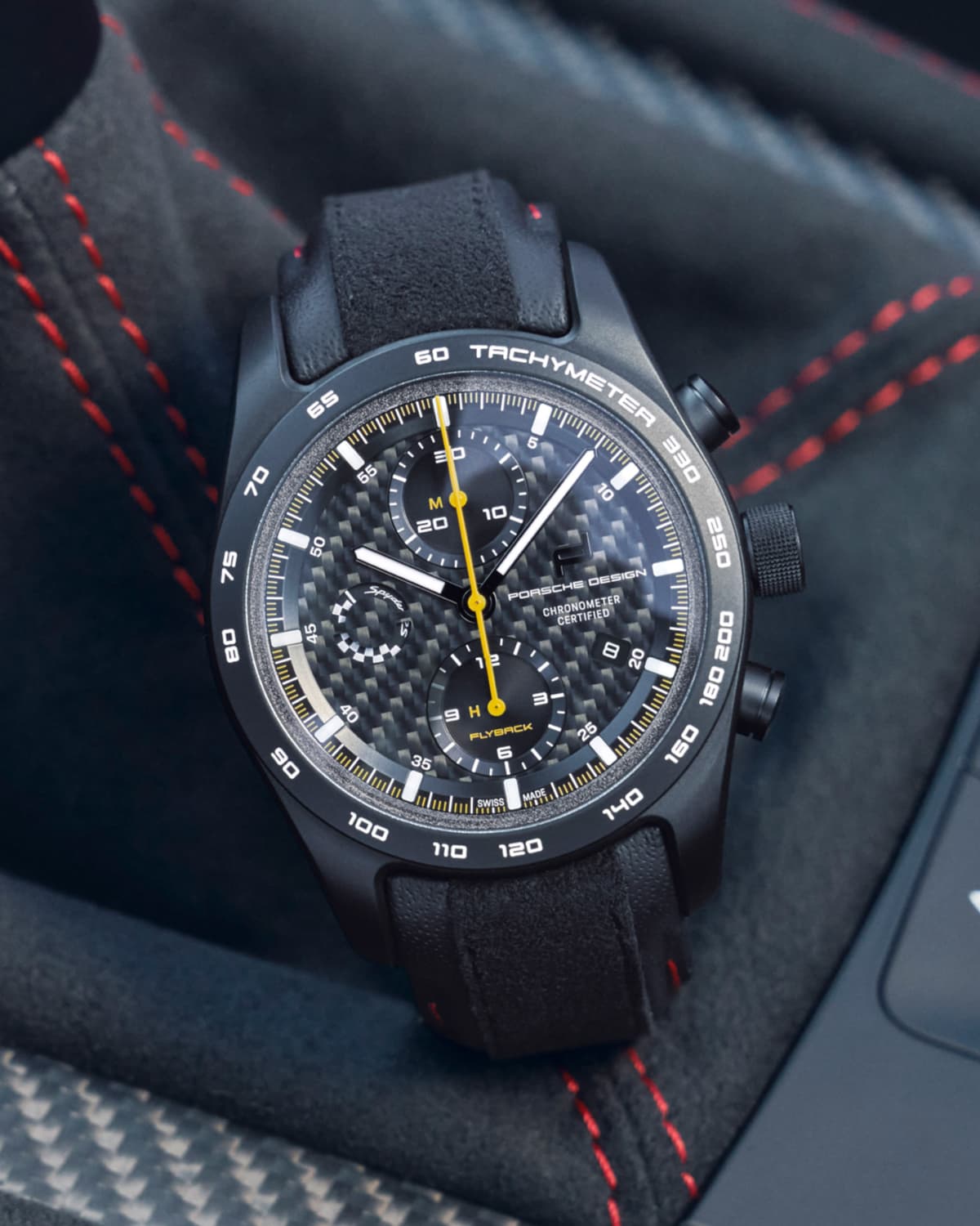 Black Porsche Design chronograph with a carbon dial and yellow seconds hand, set against a car seat with red stitching.