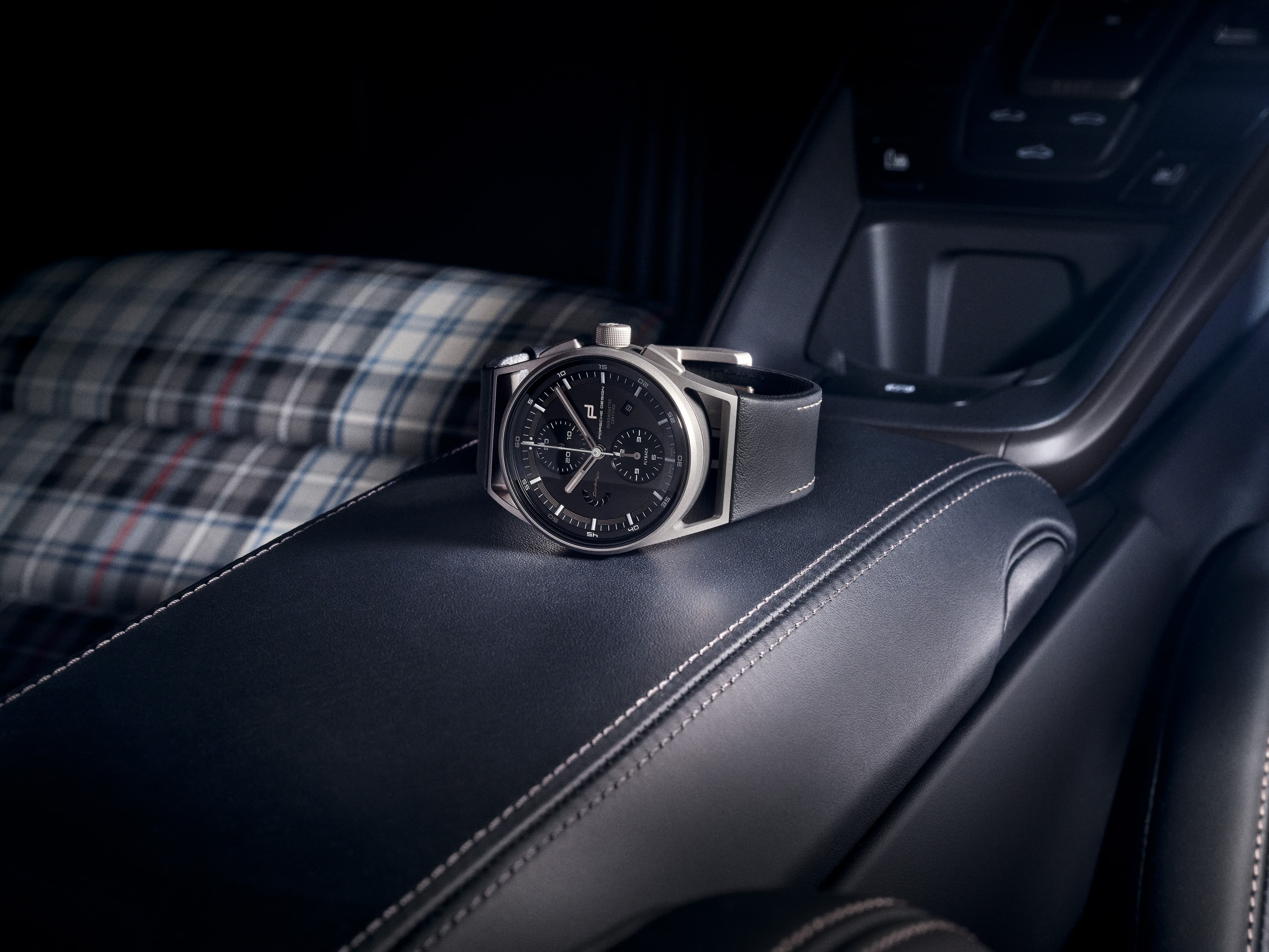 A black chronograph wristwatch with a leather strap on a black automotive leather armrest, with a checkered seat in the background.