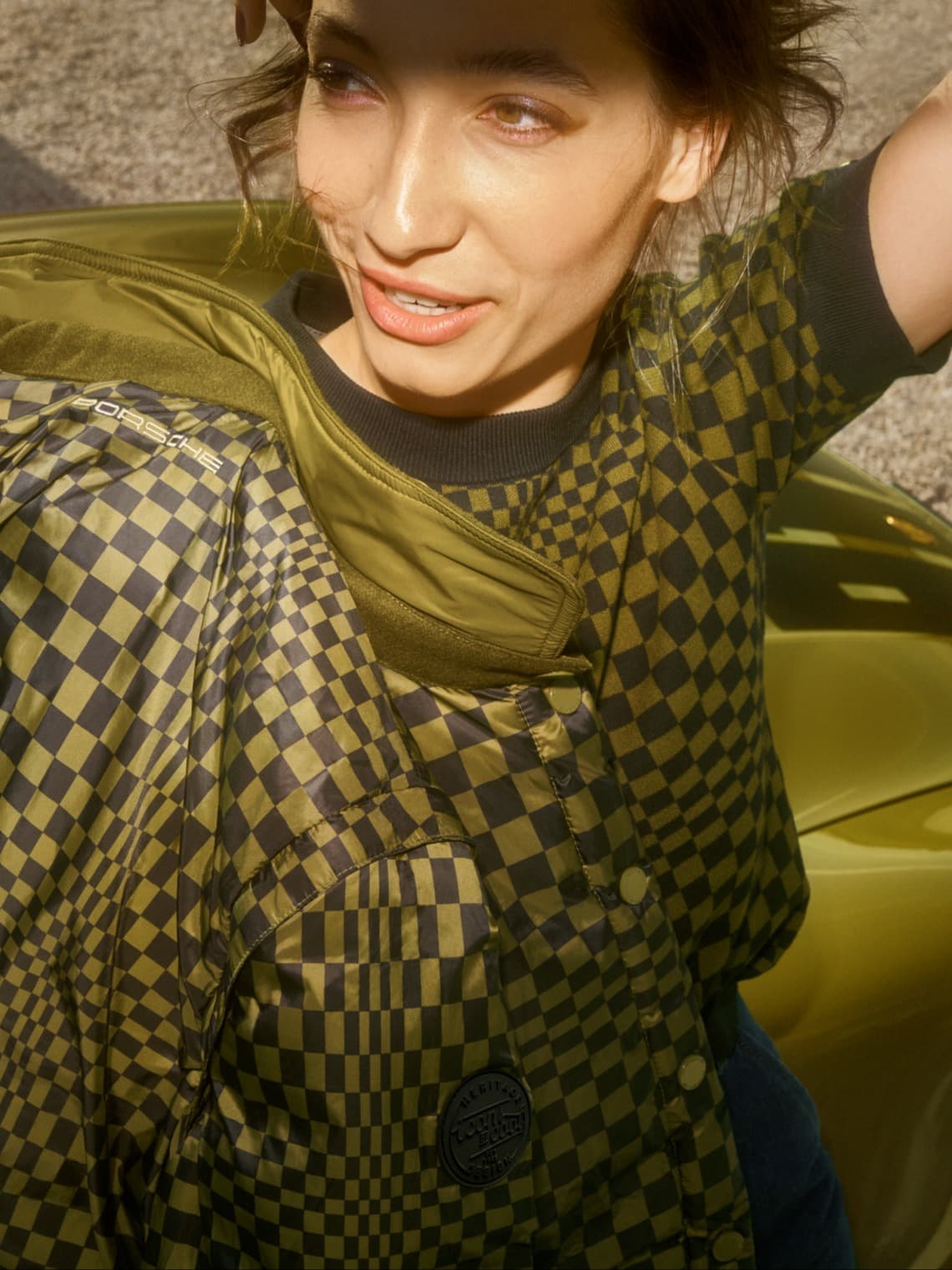 Mujer con chaqueta acolchada Porsche en verde oliva y negro con estampado de cuadros, sentada en un asiento de coche verde a la luz del sol.